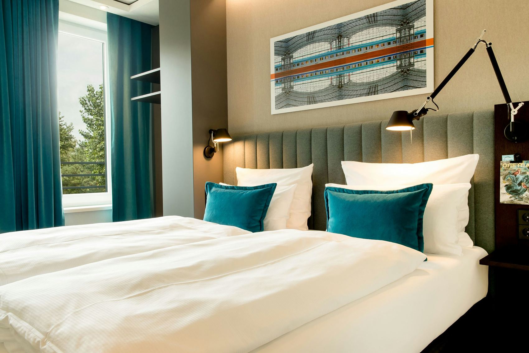 Hotel Motel One Berlin-Hauptbahnhof Modernes, helles Hotelzimmer mit großem, bequemem Doppelbett, weichen weißen Bettdecken, blauen Samtkissen und einem großen Fenster mit Blick ins Grüne. Angenehme Atmosphäre durch sanfte Beleuchtung und stilvolle Wanddekoration.