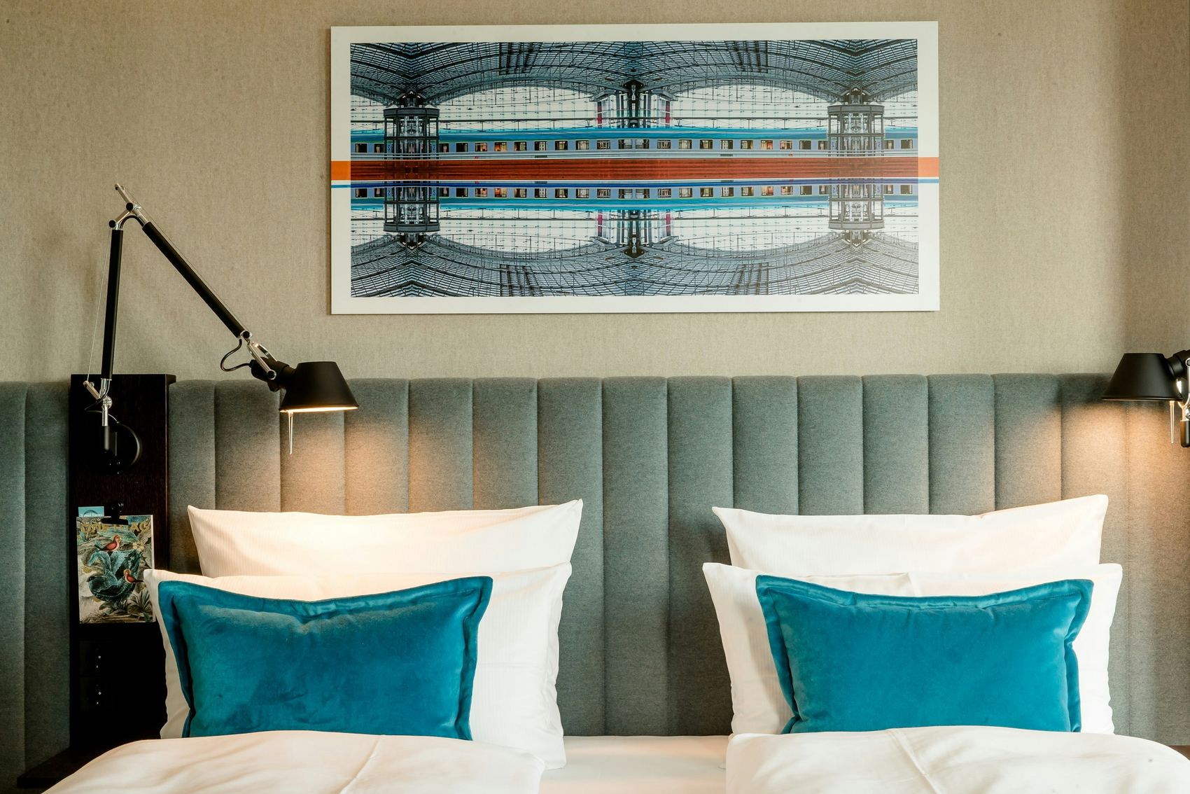Hotel Motel One Berlin-Hauptbahnhof Zwei nebeneinander stehende Betten mit weißen Bettdecken und türkisfarbenen Kissen, vor einem gepolsterten, grauen Kopfteil. Über den Betten hängt ein modernes Kunstbild mit symmetrischem Architekturmotiv. Zwei verstellbare Leselampen an den Seiten schaffen eine gemütliche Atmosphäre.