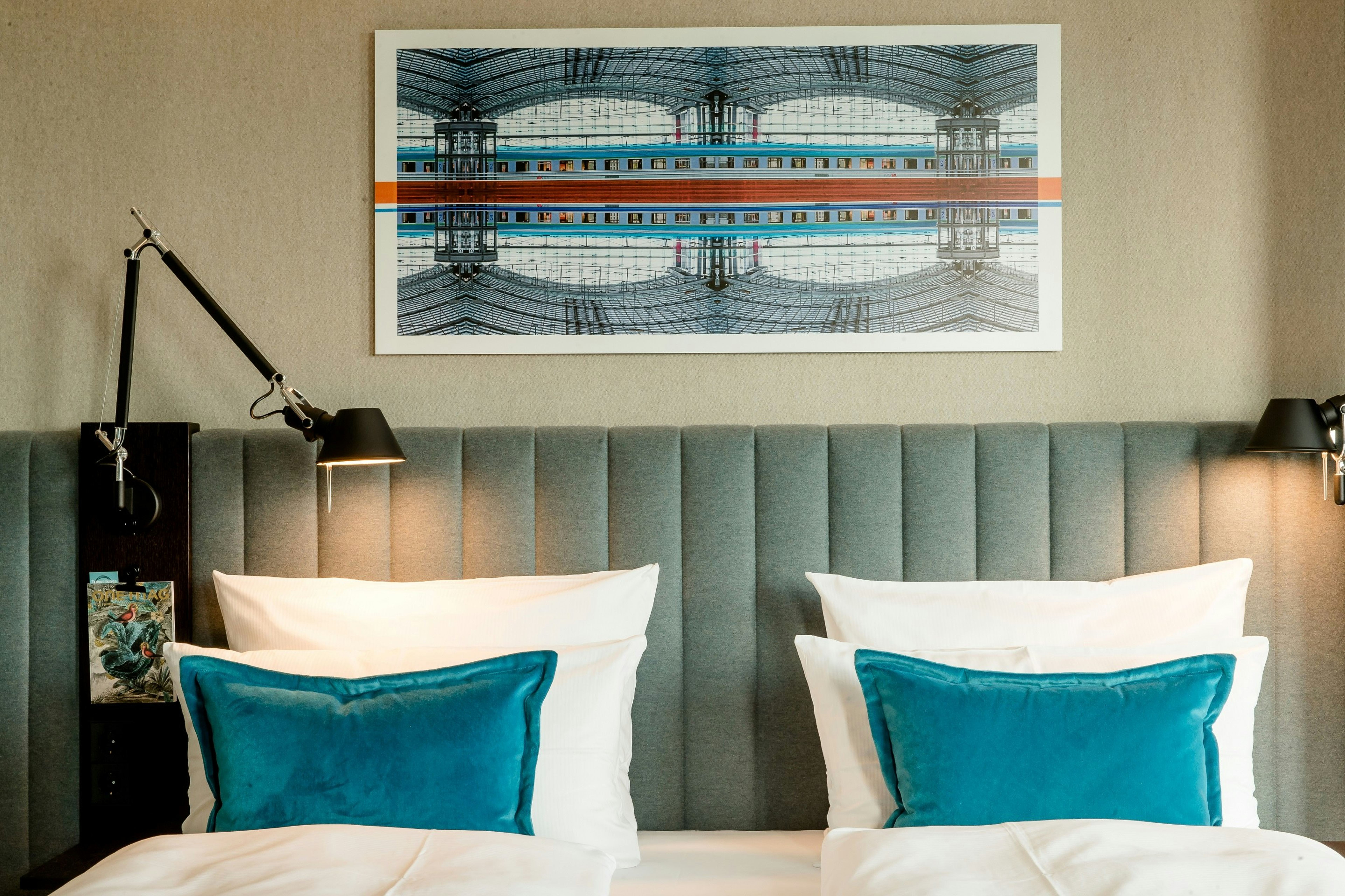 Hotel Motel One Berlin-Hauptbahnhof Zwei nebeneinander stehende Betten mit weißen Bettdecken und türkisfarbenen Kissen, vor einem gepolsterten, grauen Kopfteil. Über den Betten hängt ein modernes Kunstbild mit symmetrischem Architekturmotiv. Zwei verstellbare Leselampen an den Seiten schaffen eine gemütliche Atmosphäre. Hotel Motel One Berlin-Hauptbahnhof Zwei nebeneinander stehende Betten mit weißen Bettdecken und türkisfarbenen Kissen, vor einem gepolsterten, grauen Kopfteil. Über den Betten hängt ein modernes Kunstbild mit symmetrischem Architekturmotiv. Zwei verstellbare Leselampen an den Seiten schaffen eine gemütliche Atmosphäre.