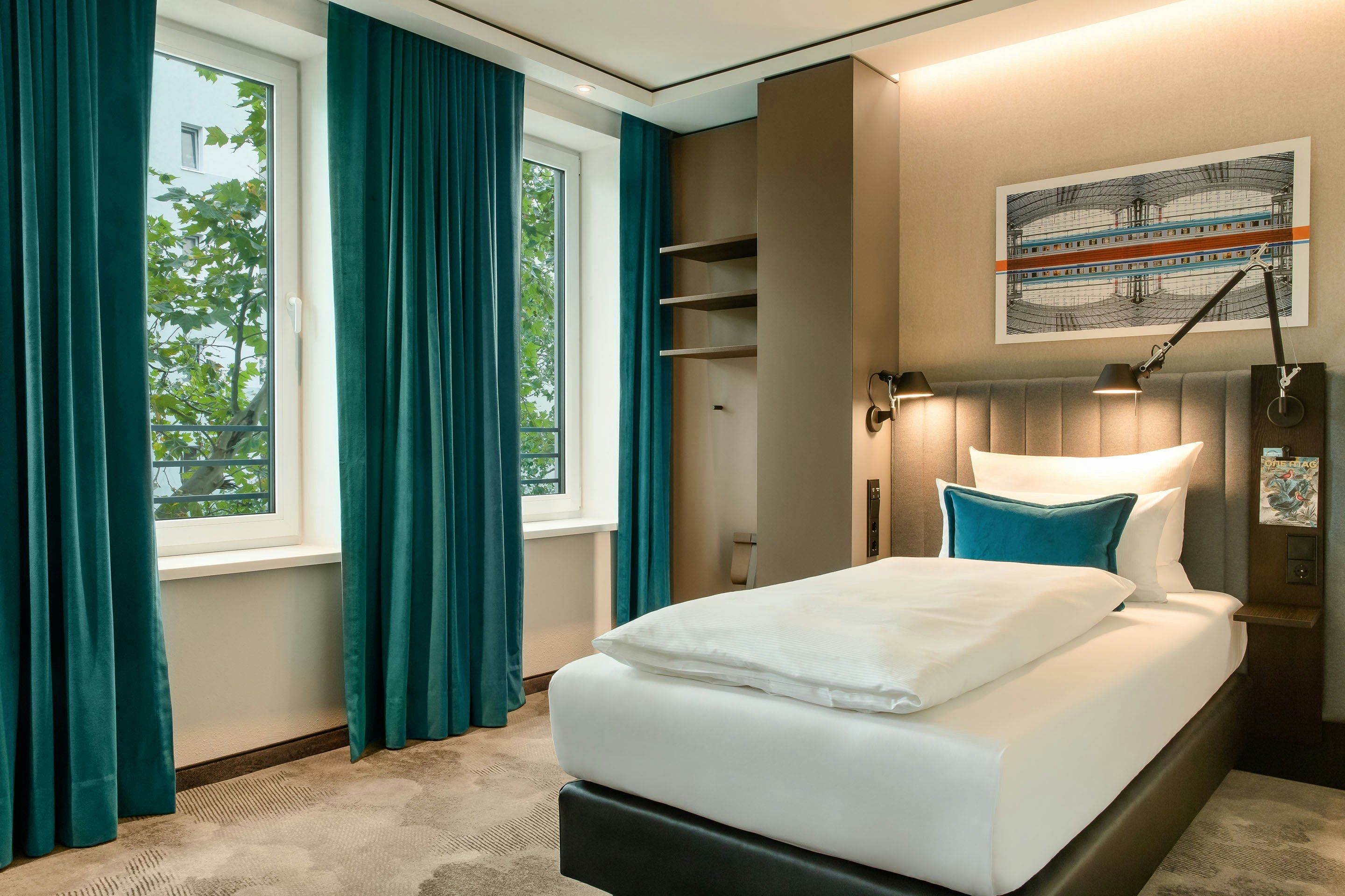 Hotel Motel One Berlin-Hauptbahnhof Modern eingerichtetes Einzelzimmer mit großem Fenster, türkisfarbenen Vorhängen, gemütlichem Bett und warmem Licht für entspannte Nächte.