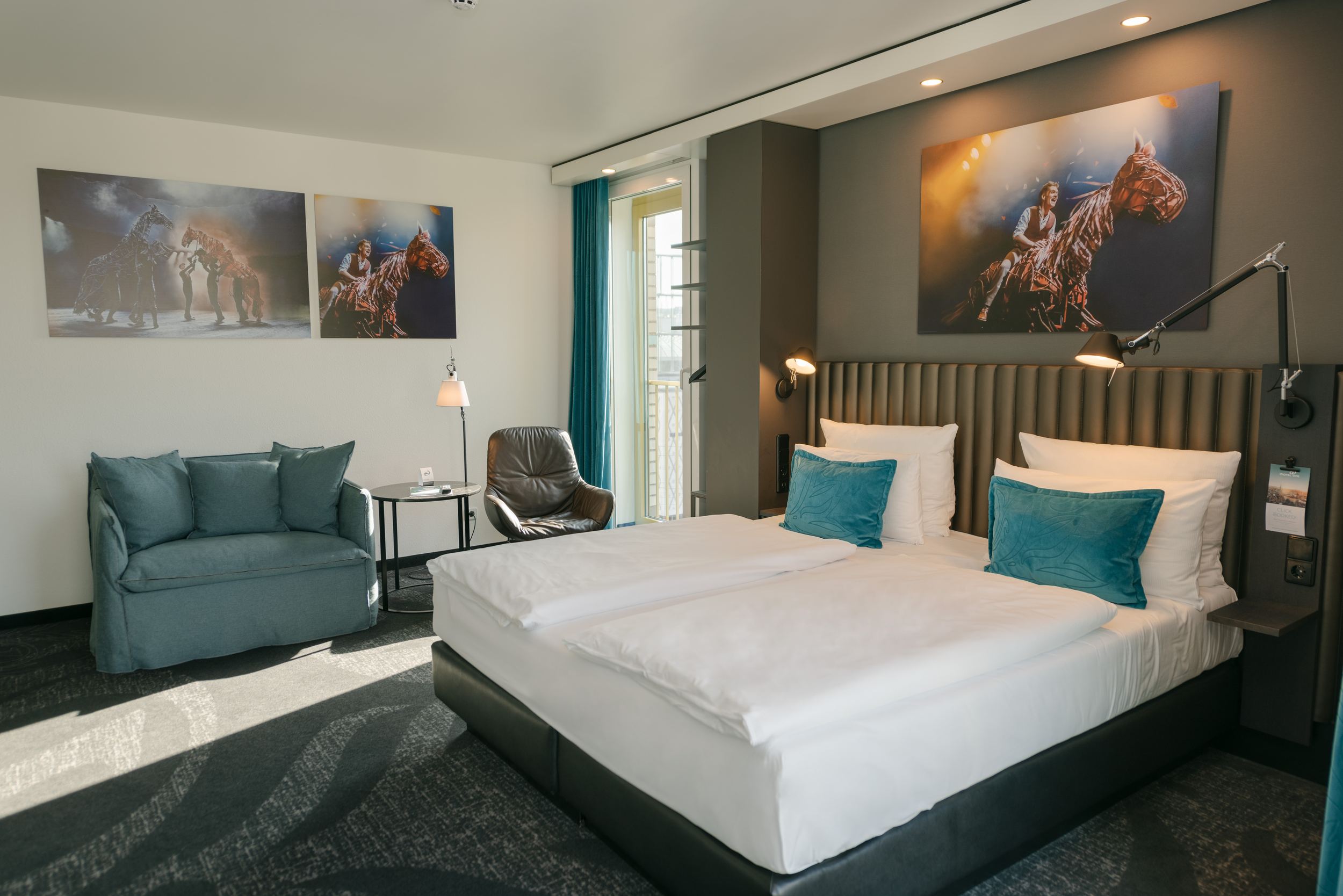 Hotel Berlin Ku’damm Motel One helles Zimmer mit Doppelbett, Sofa, Sessel und Beistelltisch sowie bodentiefes Fenster mit direkter Sonneneinstrahlung