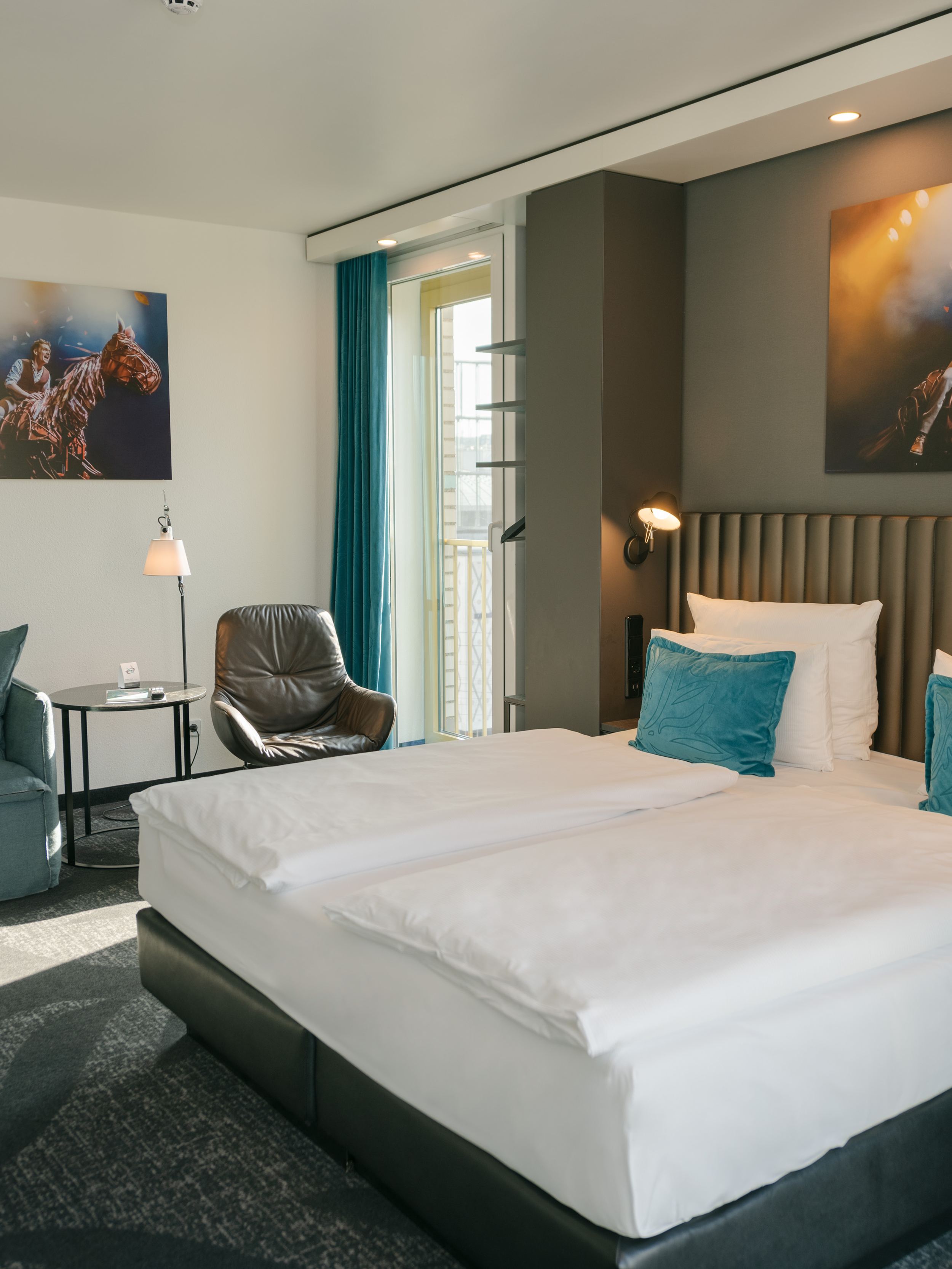 Hotel Berlin Ku’damm Motel One helles Zimmer mit Doppelbett, Sofa, Sessel und Beistelltisch sowie bodentiefes Fenster mit direkter Sonneneinstrahlung