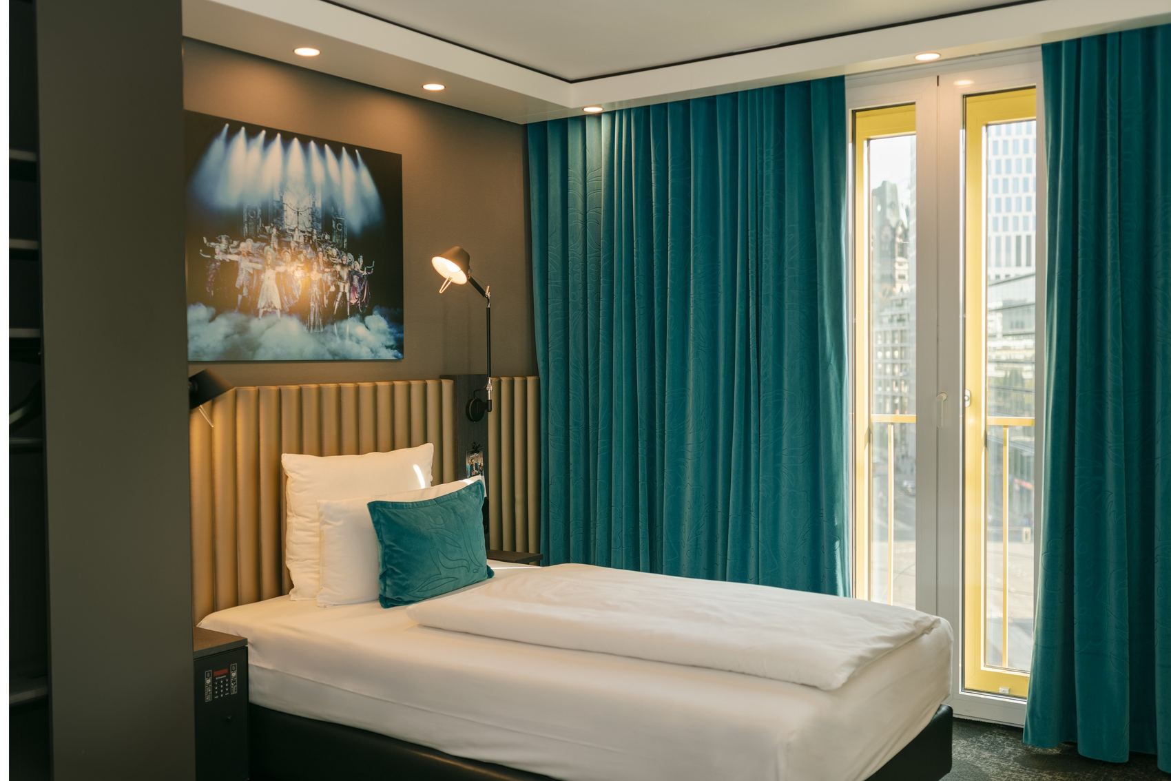 Modernes Hotelzimmer mit Einzelbett, elegantem Kopfteil und Akzentkissen in Blau. Große Fenster mit blauen Vorhängen bieten Tageslicht und Stadtblick. Gedimmte Beleuchtung und stilvolles Wandbild schaffen eine entspannte Atmosphäre.