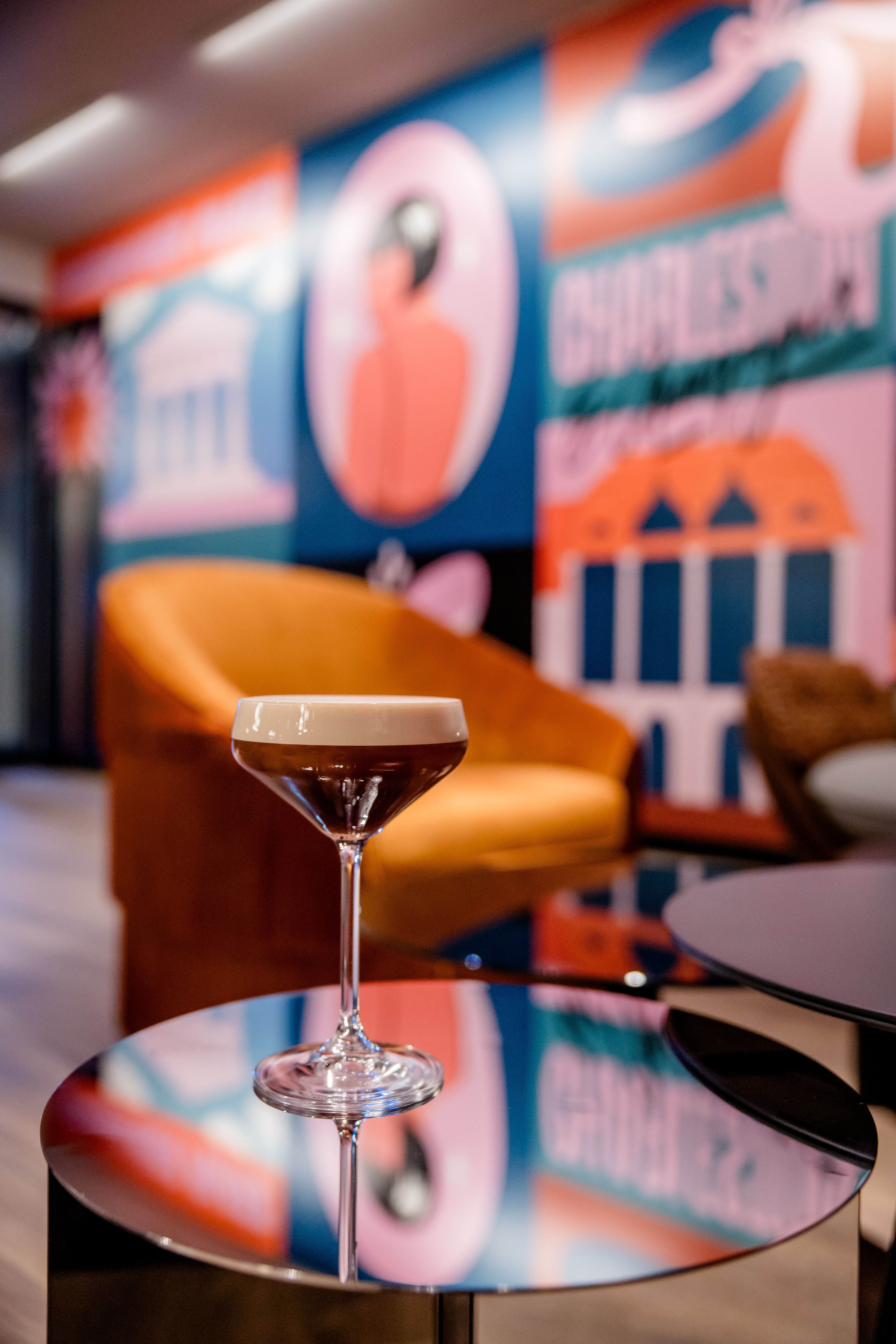 Hotel Berlin-Potsdamer Platz Motel One Cocktailglas mit Schaum auf einem spiegelnden Tisch, im Hintergrund eine gemütliche orangefarbene Sitzgelegenheit und bunte, moderne Wandkunst mit stilisierten Figuren und Gebäuden.