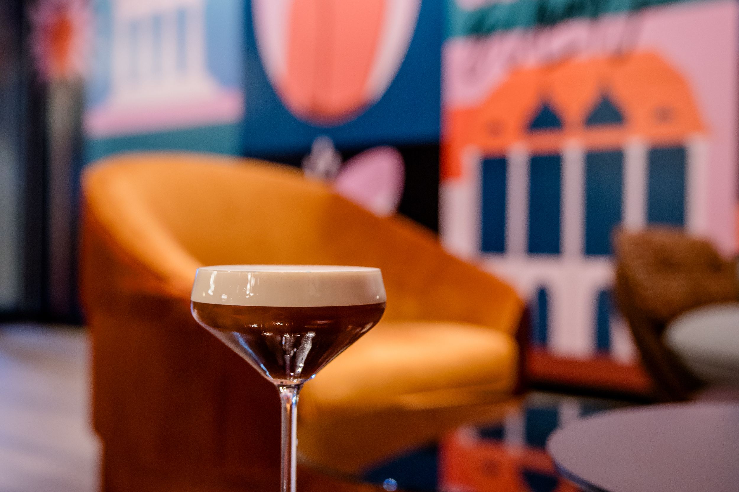 Hotel Berlin-Potsdamer Platz Motel One Cocktailglas mit Schaum auf einem spiegelnden Tisch, im Hintergrund eine gemütliche orangefarbene Sitzgelegenheit und bunte, moderne Wandkunst mit stilisierten Figuren und Gebäuden.
