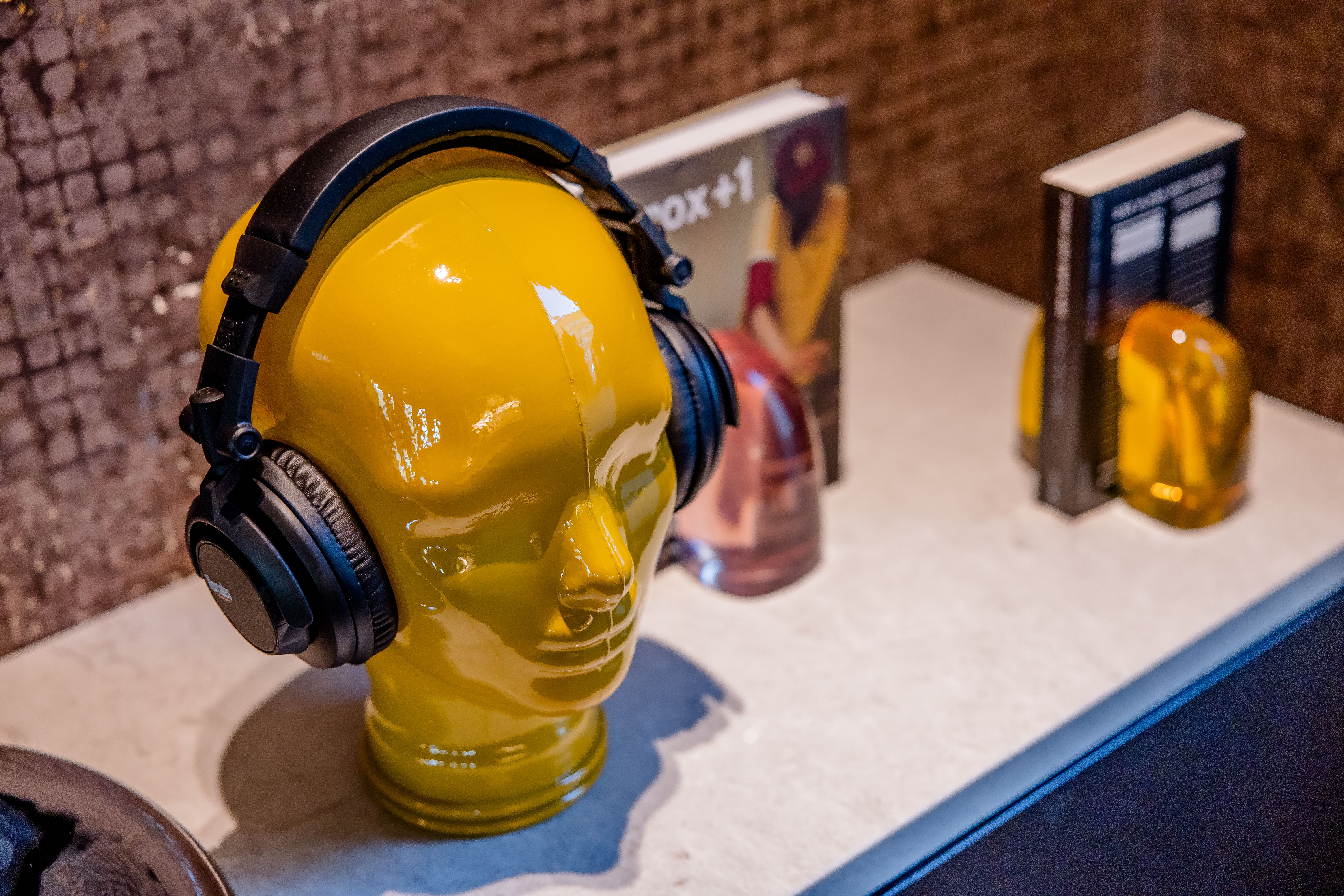 Hotel Berlin-Potsdamer Platz Motel One und gelber Dekokopf mit schwarzen Kopfhörern steht auf einer weißen Oberfläche vor einer Wand, daneben Bücher und weitere kleine Dekorationsgegenstände.