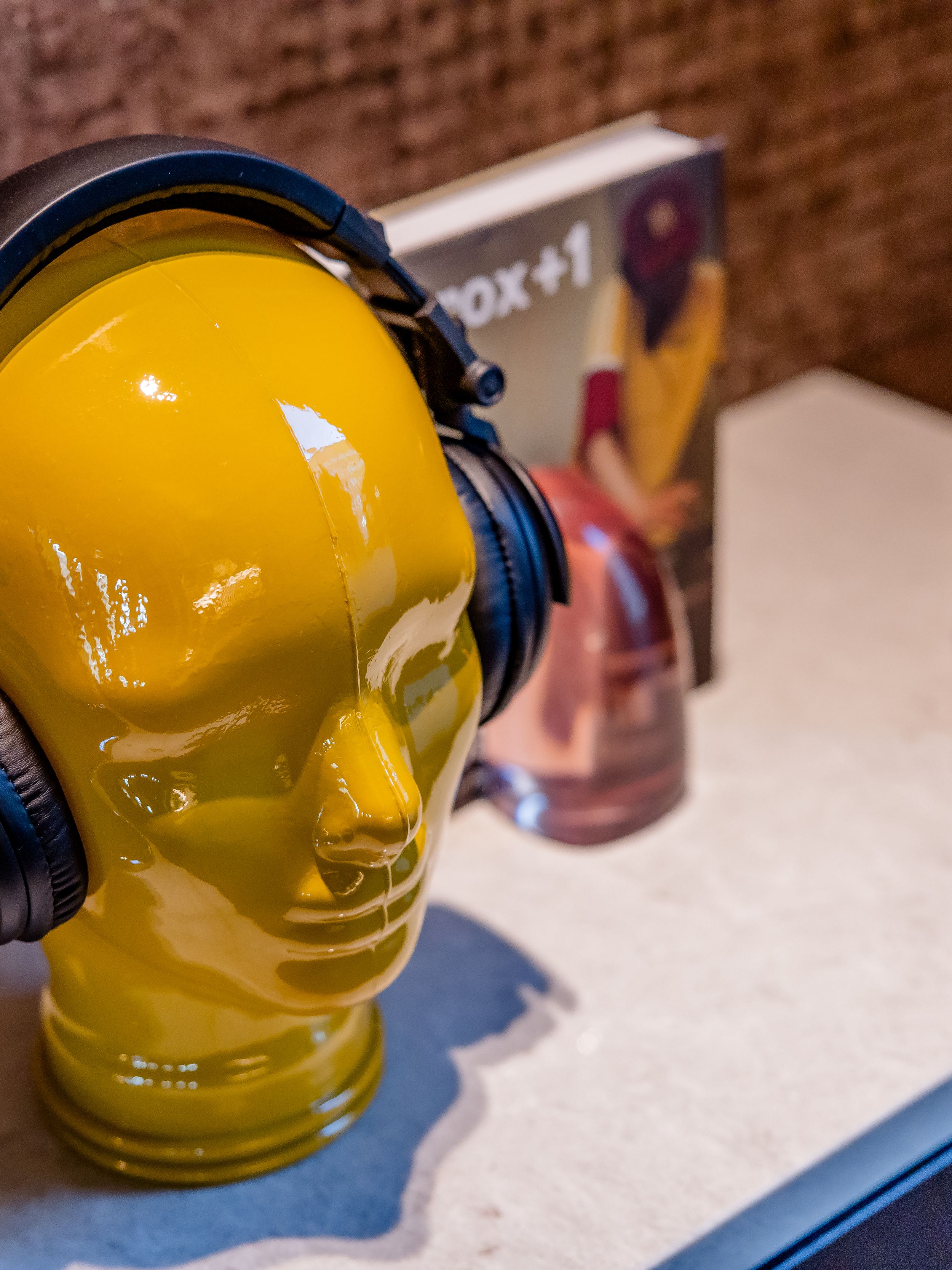 Hotel Berlin-Potsdamer Platz Motel One und gelber Dekokopf mit schwarzen Kopfhörern steht auf einer weißen Oberfläche vor einer Wand, daneben Bücher und weitere kleine Dekorationsgegenstände.