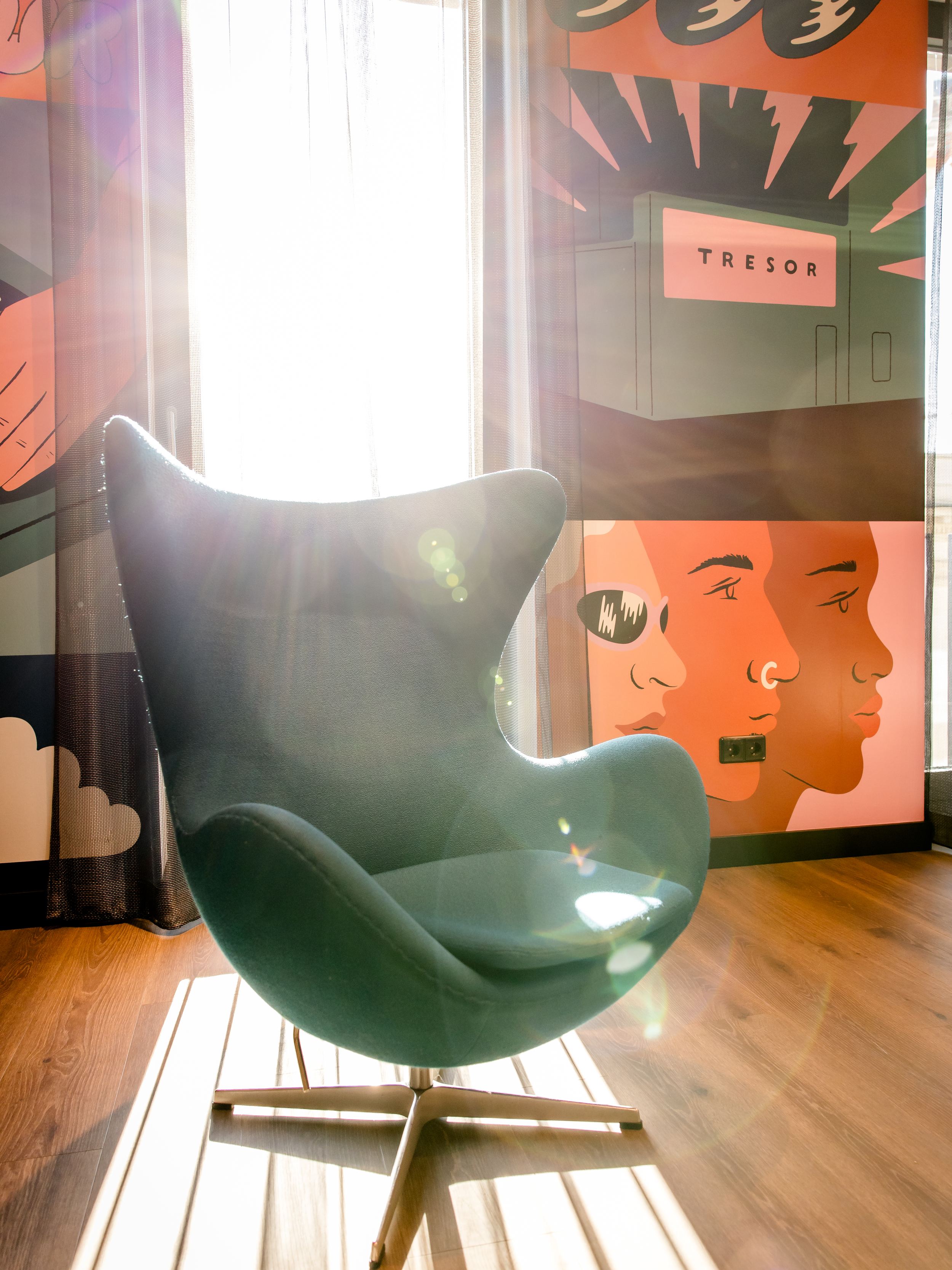 Hotel Berlin-Potsdamer Platz Motel One Moderner, dunkelgrüner Egg Chair vor großem Fenster mit Sonnenlicht. Farbige Wandkunst mit stilisierten Gesichtern und Schallplatten im Hintergrund.