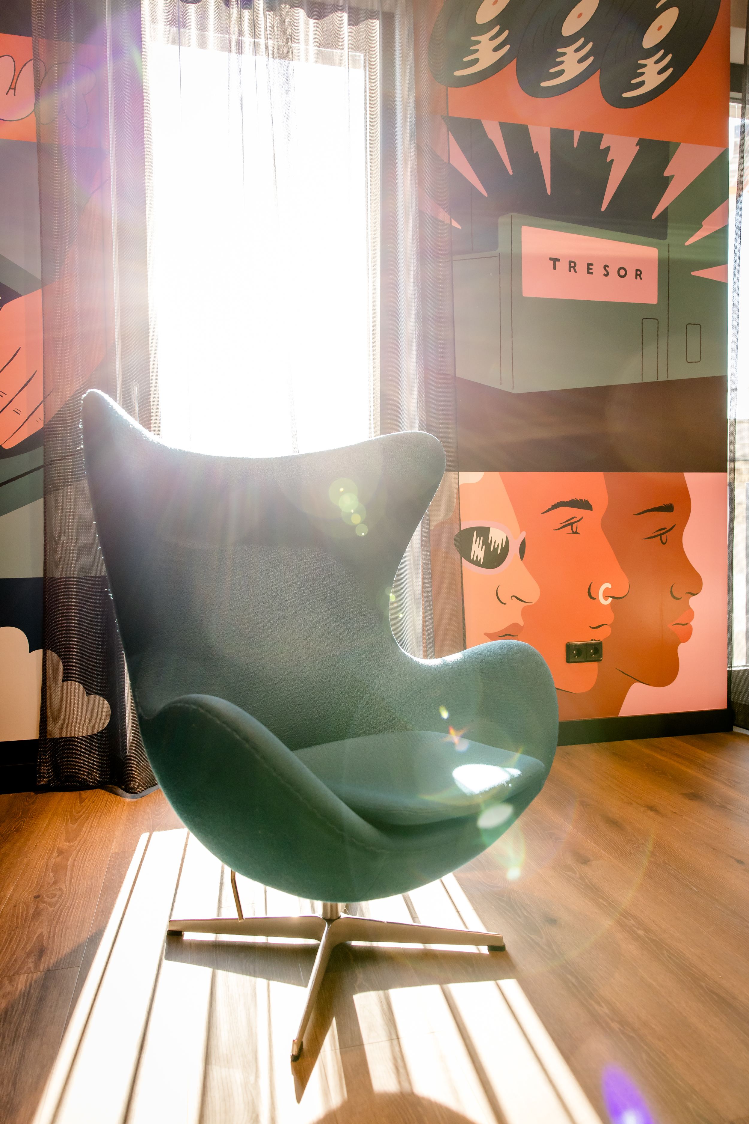 Hotel Berlin-Potsdamer Platz Motel One Moderner, dunkelgrüner Egg Chair vor großem Fenster mit Sonnenlicht. Farbige Wandkunst mit stilisierten Gesichtern und Schallplatten im Hintergrund.