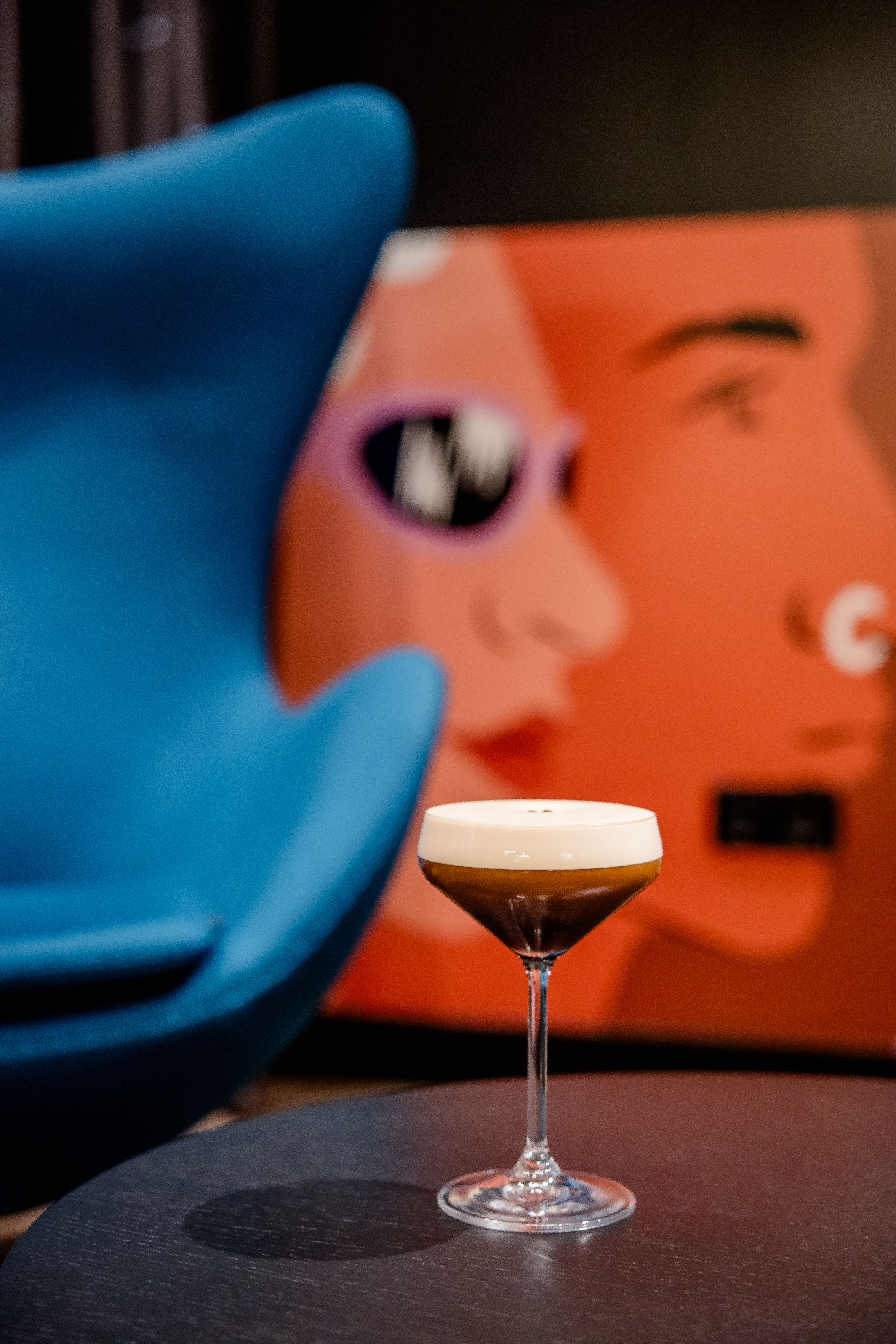 Hotel Berlin-Potsdamer Platz Motel One Cocktailglas mit schaumigem Getränk auf einem Tisch, im Hintergrund unscharfe, moderne Wandkunst mit stilisierten Gesichtern und ein blauer Sessel.