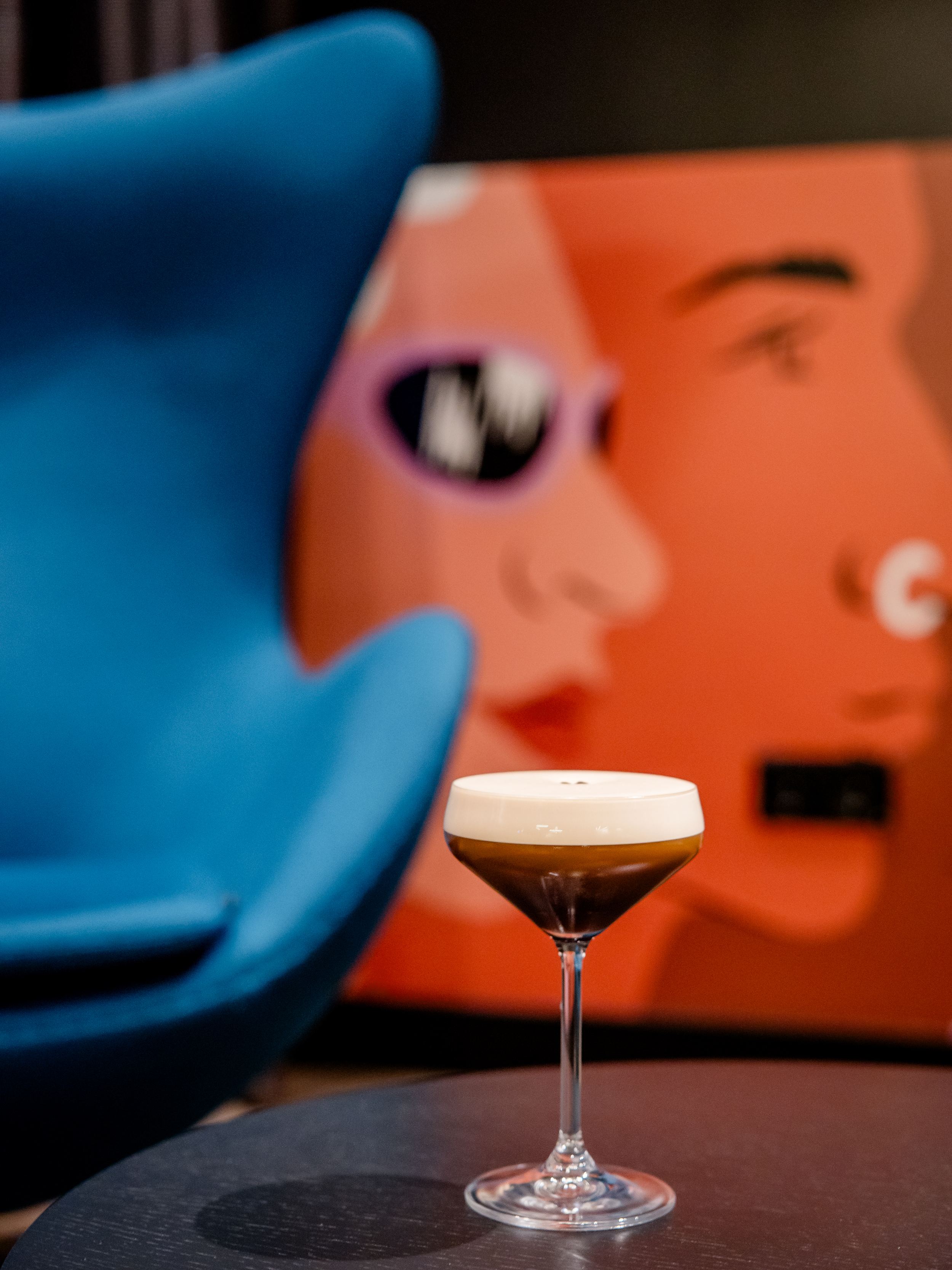 Hotel Berlin-Potsdamer Platz Motel One Cocktailglas mit schaumigem Getränk auf einem Tisch, im Hintergrund unscharfe, moderne Wandkunst mit stilisierten Gesichtern und ein blauer Sessel.