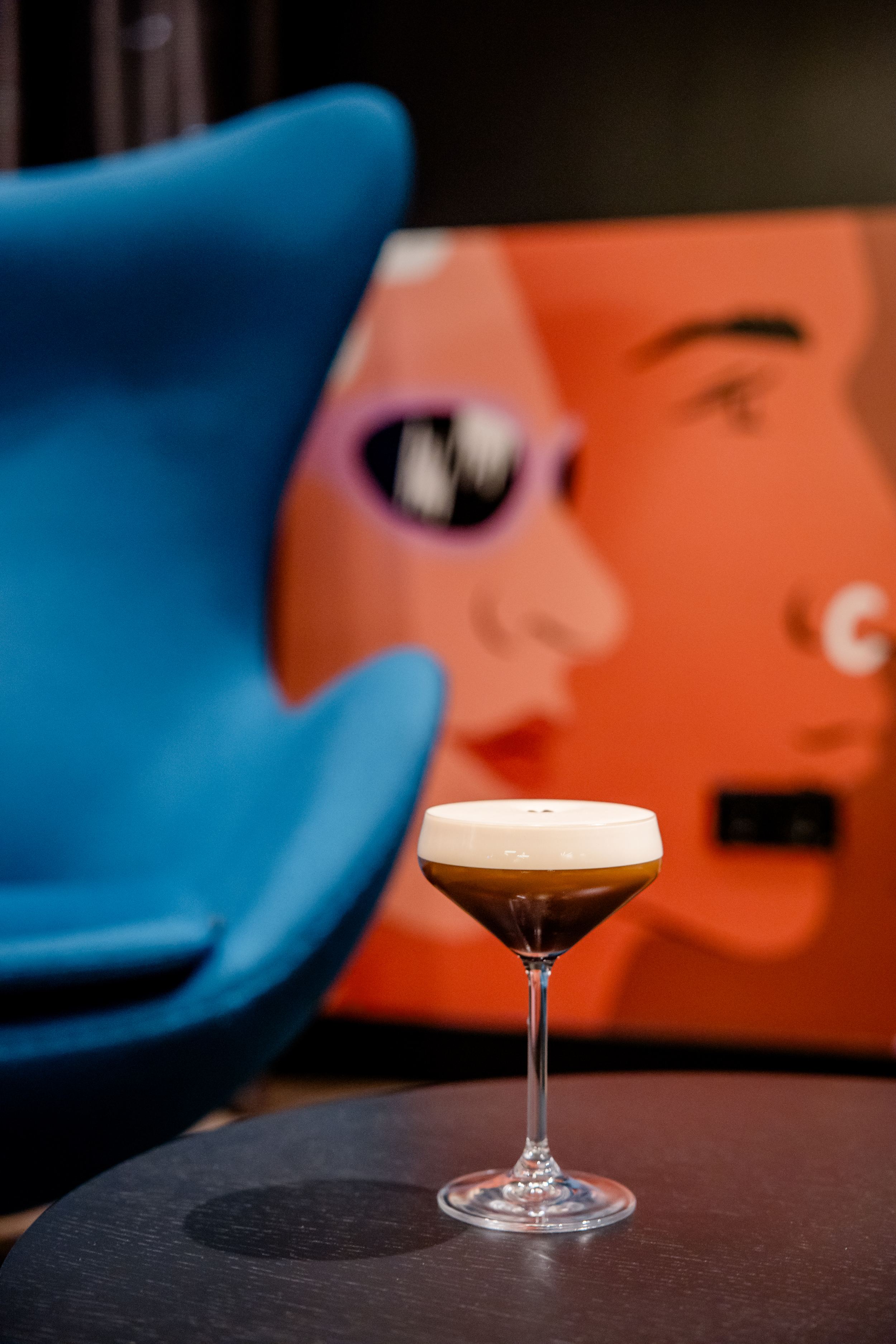 Hotel Berlin-Potsdamer Platz Motel One Cocktailglas mit schaumigem Getränk auf einem Tisch, im Hintergrund unscharfe, moderne Wandkunst mit stilisierten Gesichtern und ein blauer Sessel.