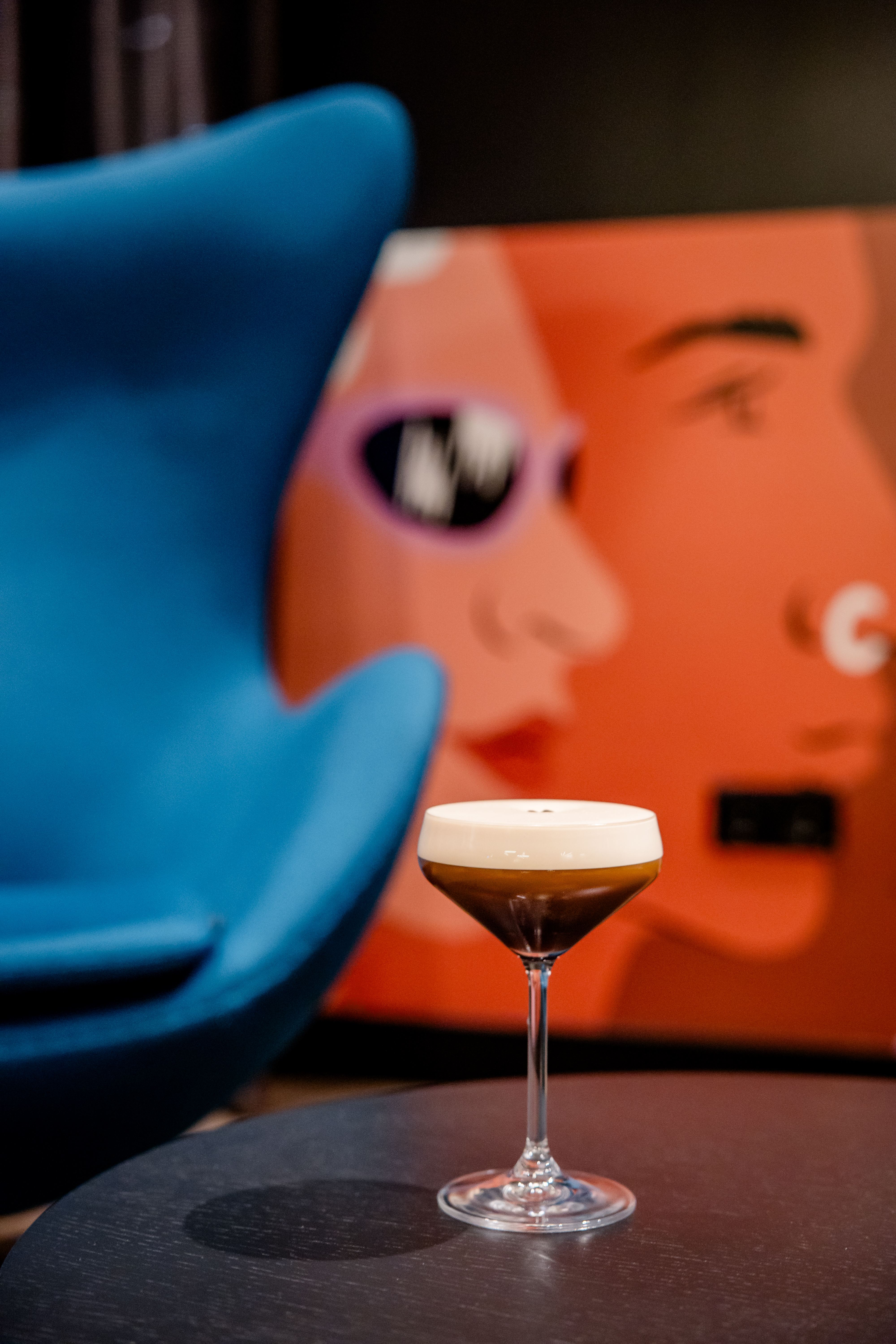 Hotel Berlin-Potsdamer Platz Motel One Cocktailglas mit schaumigem Getränk auf einem Tisch, im Hintergrund unscharfe, moderne Wandkunst mit stilisierten Gesichtern und ein blauer Sessel. Hotel Berlin-Potsdamer Platz Motel One Cocktailglas mit schaumigem Getränk auf einem Tisch, im Hintergrund unscharfe, moderne Wandkunst mit stilisierten Gesichtern und ein blauer Sessel.
