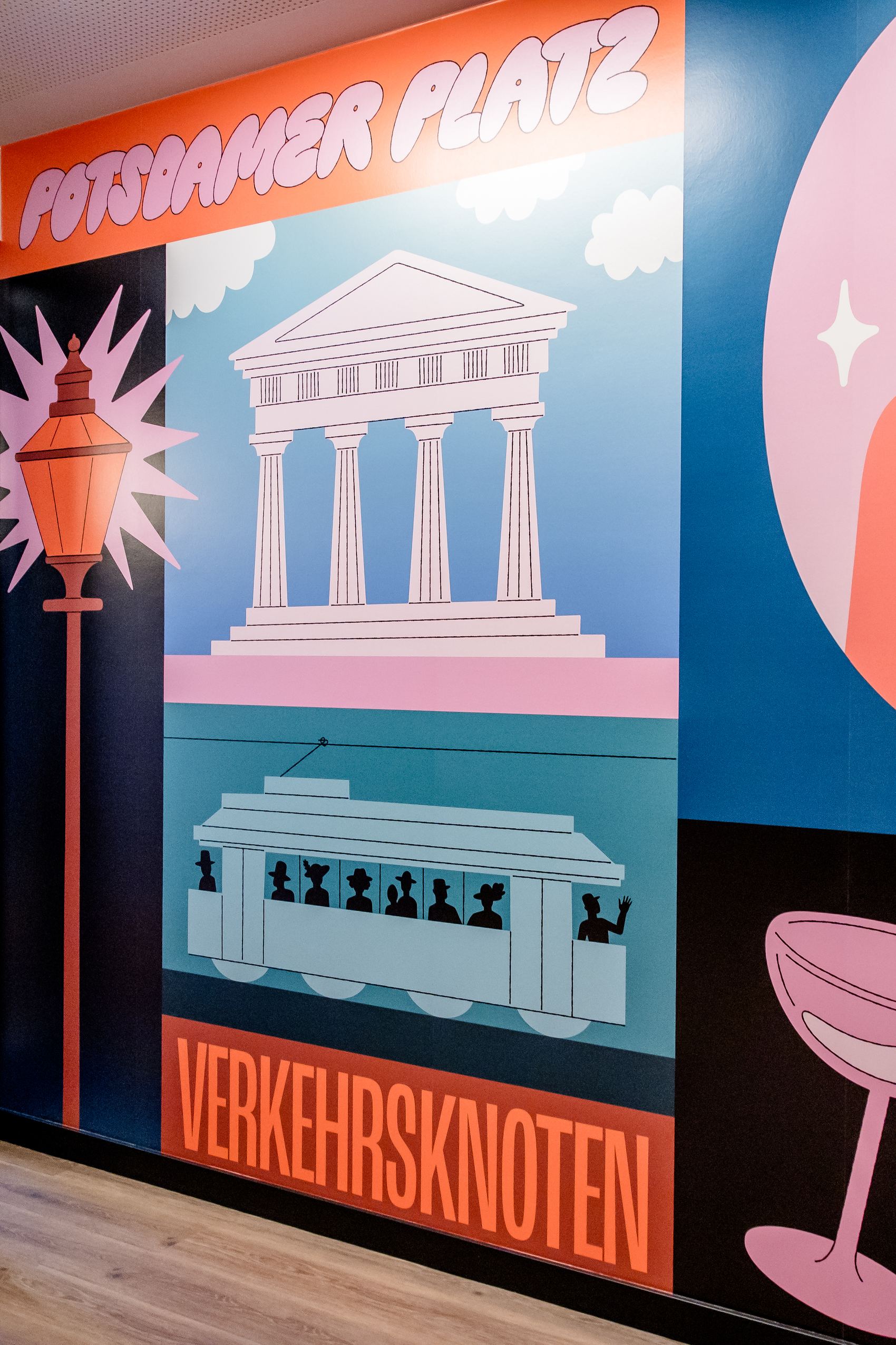 Hotel Berlin-Potsdamer Platz Motel One bunte Wandgestaltung mit Illustrationen eines Tempels, einer Straßenlampe, einer Straßenbahn, einem stilisierten Porträt einer Frau und zwei anstoßenden Sektgläsern, daneben Eingang zu einer Bar mit blauem Neonlicht „One Bar“.