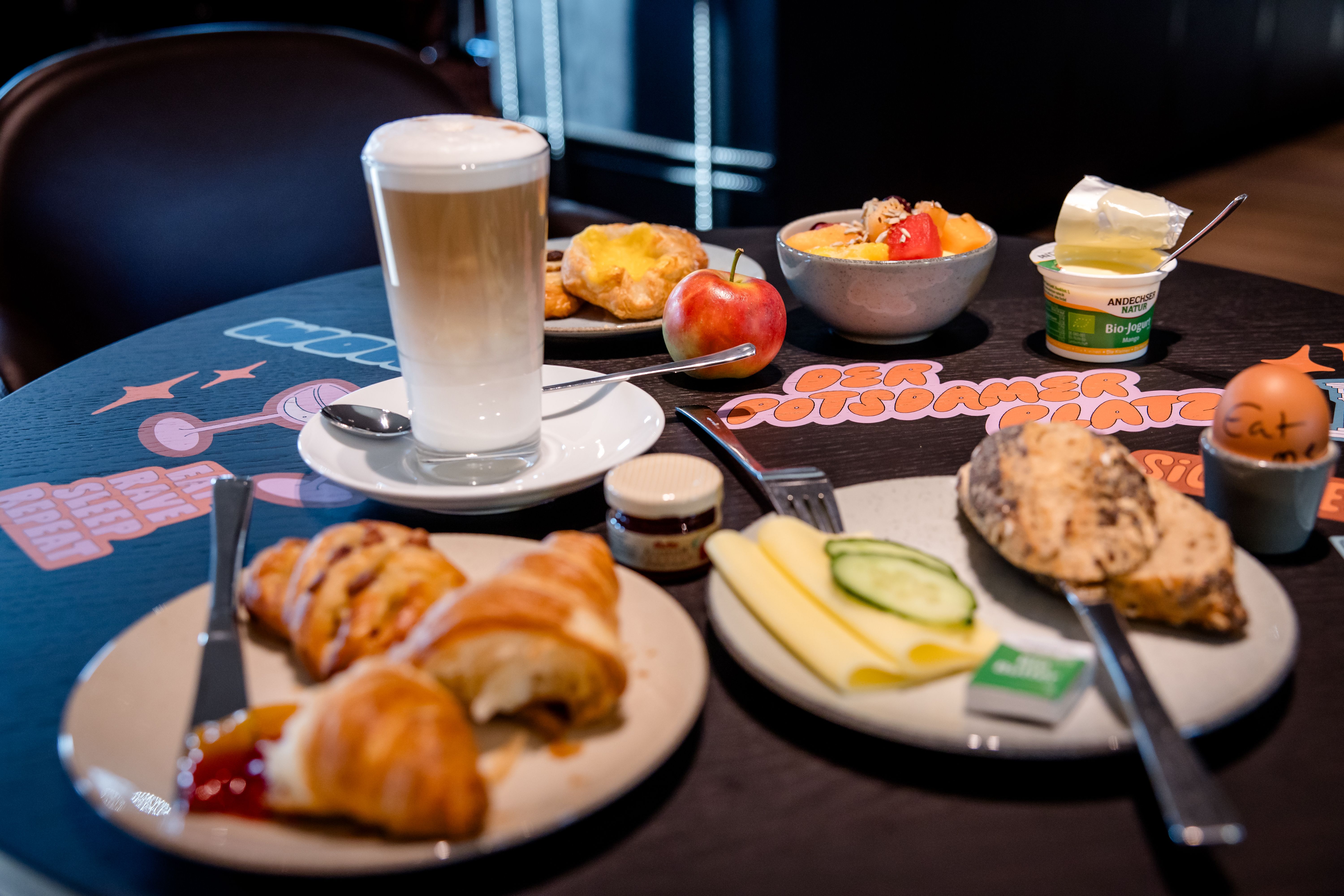 Hotel Berlin-Potsdamer Platz Motel One Frühstücksteller mit Croissants, Käse, Gurkenscheiben, ein weichgekochtes Ei, Apfel, Joghurt, Müsli und ein Glas Latte Macchiato auf einem Tisch in entspannter Atmosphäre.