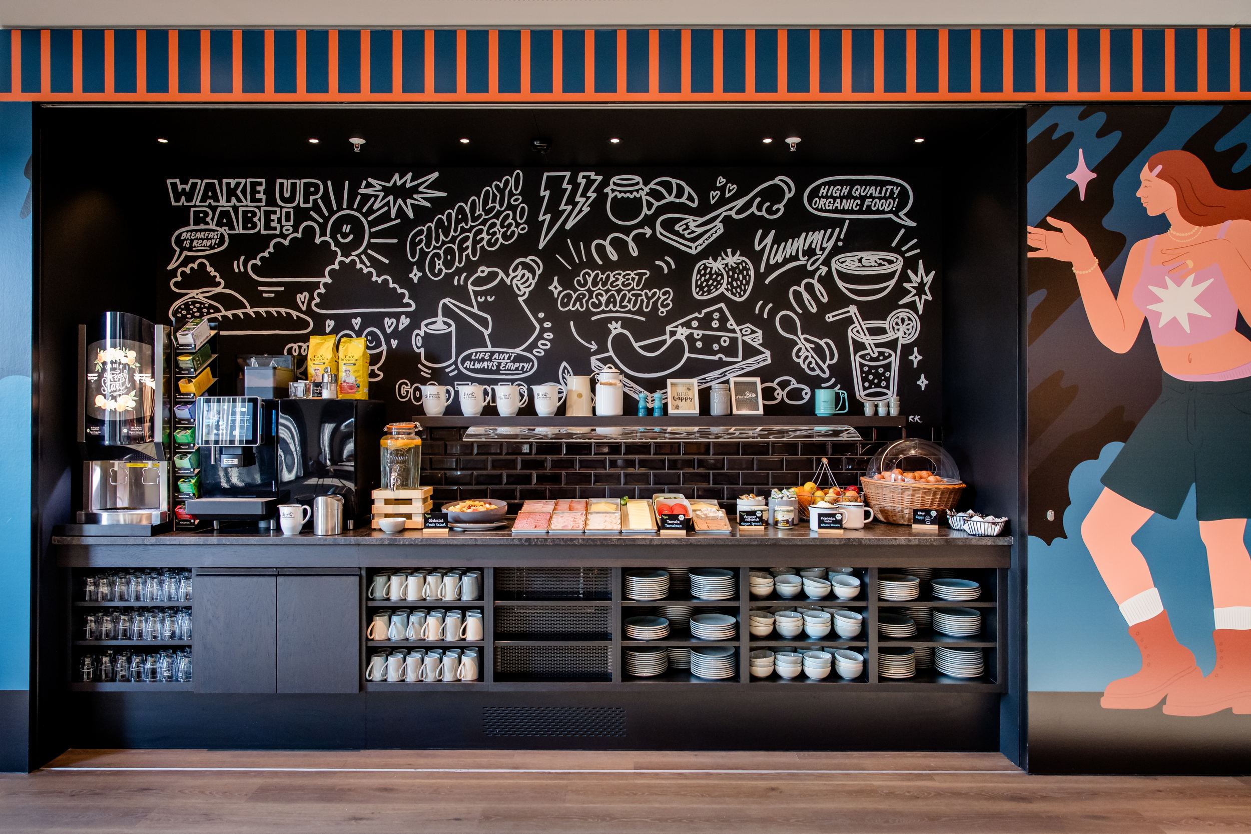 Hotel Berlin-Potsdamer Platz Motel One Selbstbedienungsbereich mit Kaffeemaschine, Teekannen, Frühstücksaufschnitten und Geschirr, vor einer Wand mit beleuchteten, handgezeichneten Motiven und Kaffee-Symbolen.