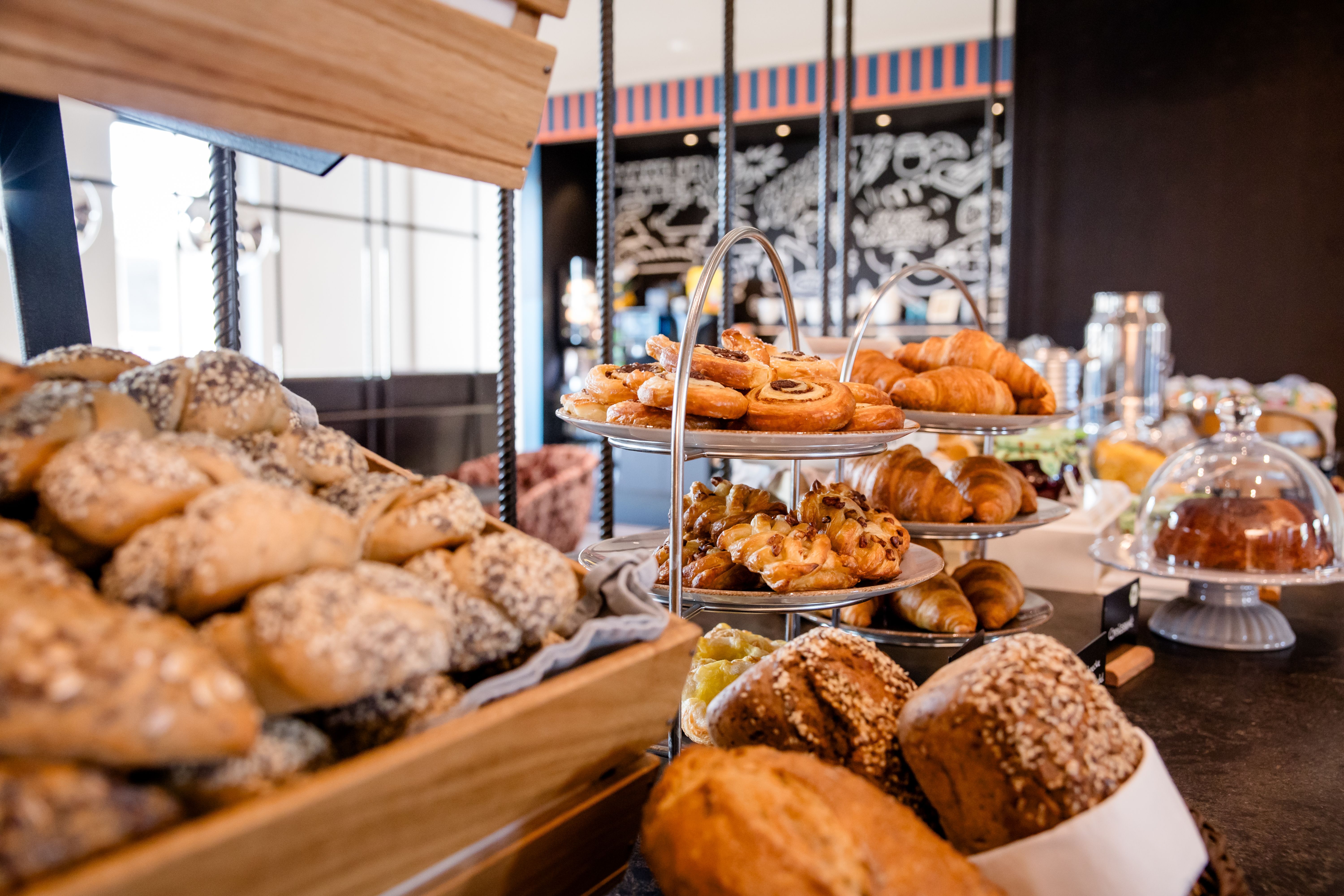 Hotel Berlin-Potsdamer Platz Motel One Vielfältiges Frühstücksbuffet mit frisch gebackenen Brötchen, Croissants, süßen und herzhaften Backwaren, ansprechend angerichtet auf Holz- und Metallständern in einem modernen Restaurant.