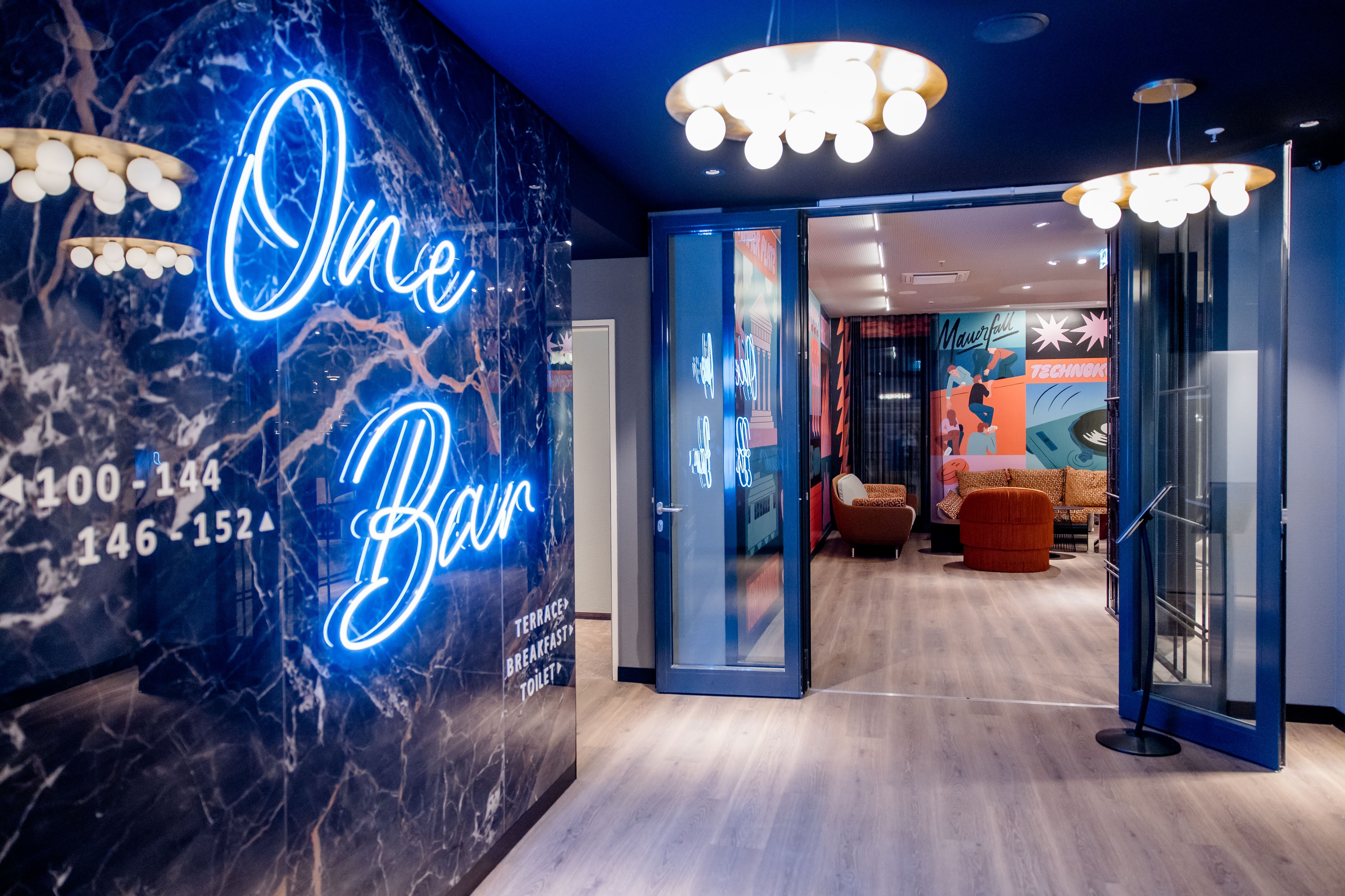 Hotel Berlin-Potsdamer Platz Motel One Eingangsbereich einer stilvollen Hotelbar mit blauem Neonlicht „One Bar“, eleganter Beleuchtung und moderner Lounge mit bunten Wandbildern, gemütlichen Sesseln und einladender Atmosphäre.