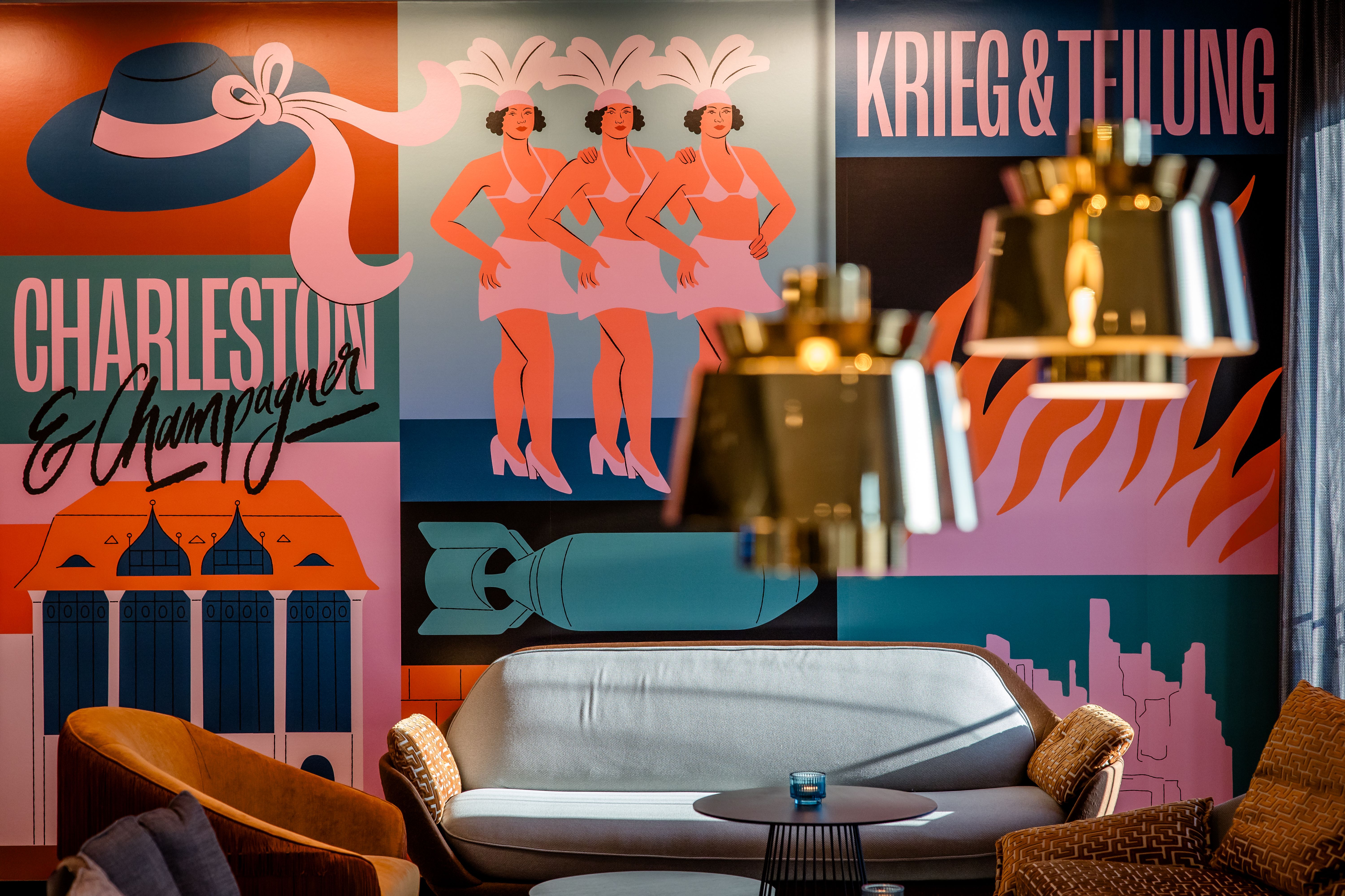 Hotel Berlin-Potsdamer Platz Motel One Bunte Wandgestaltung mit tanzenden Charleston-Frauen, Kriegssymbolen und der Schrift „Krieg & Teilung“. Gemütliche Sitzgruppe mit Sofas und Leuchten im Vordergrund.