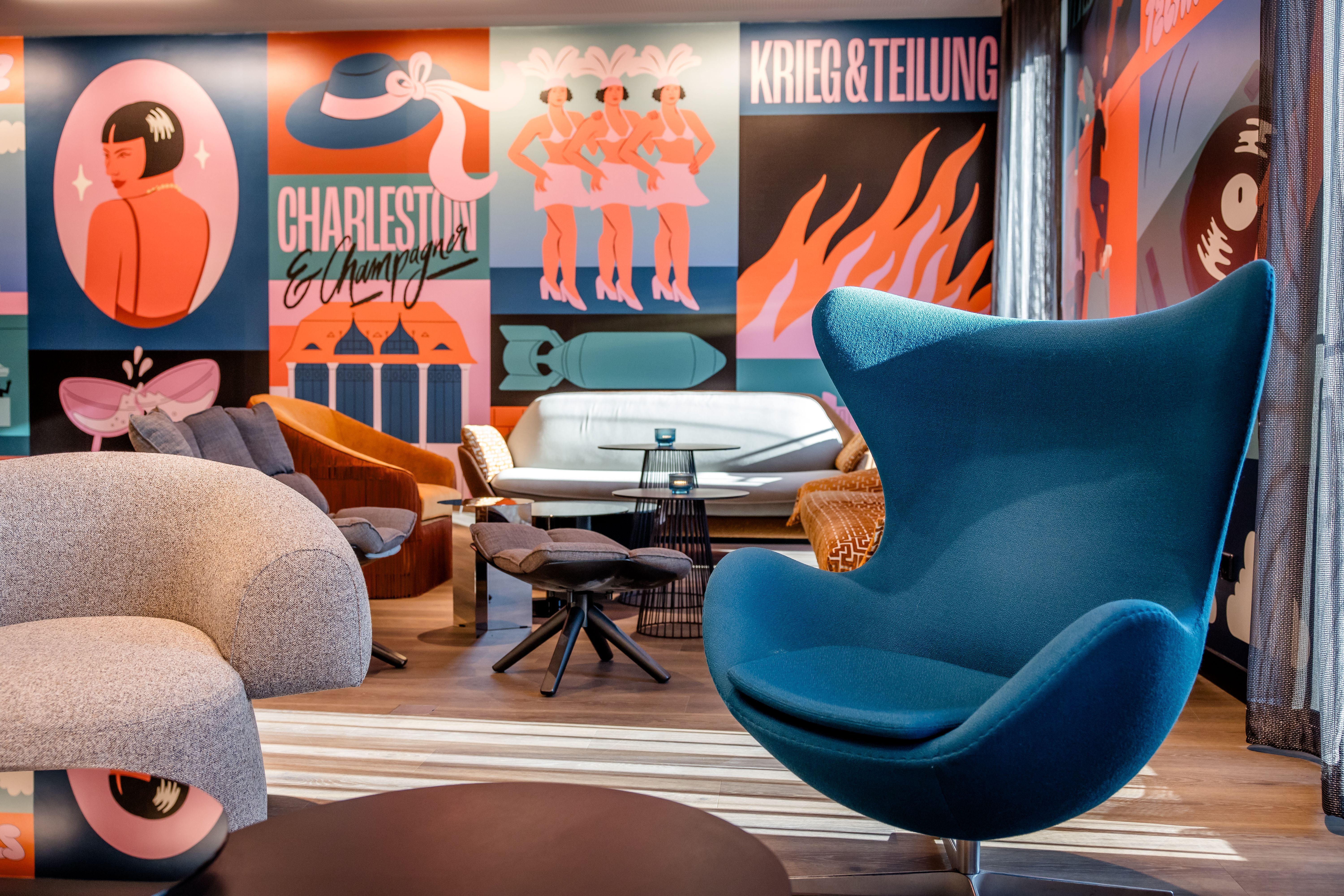 Hotel Berlin-Potsdamer Platz mit One Lounge mit modernen Sesseln und Sofas vor farbenfrohen Wandbildern im Retro-Stil, die lebendige Motive und Schriftzüge zeigen. Helle, einladende Atmosphäre mit stilvollem Design und bequemen Sitzgelegenheiten für entspannte Momente.