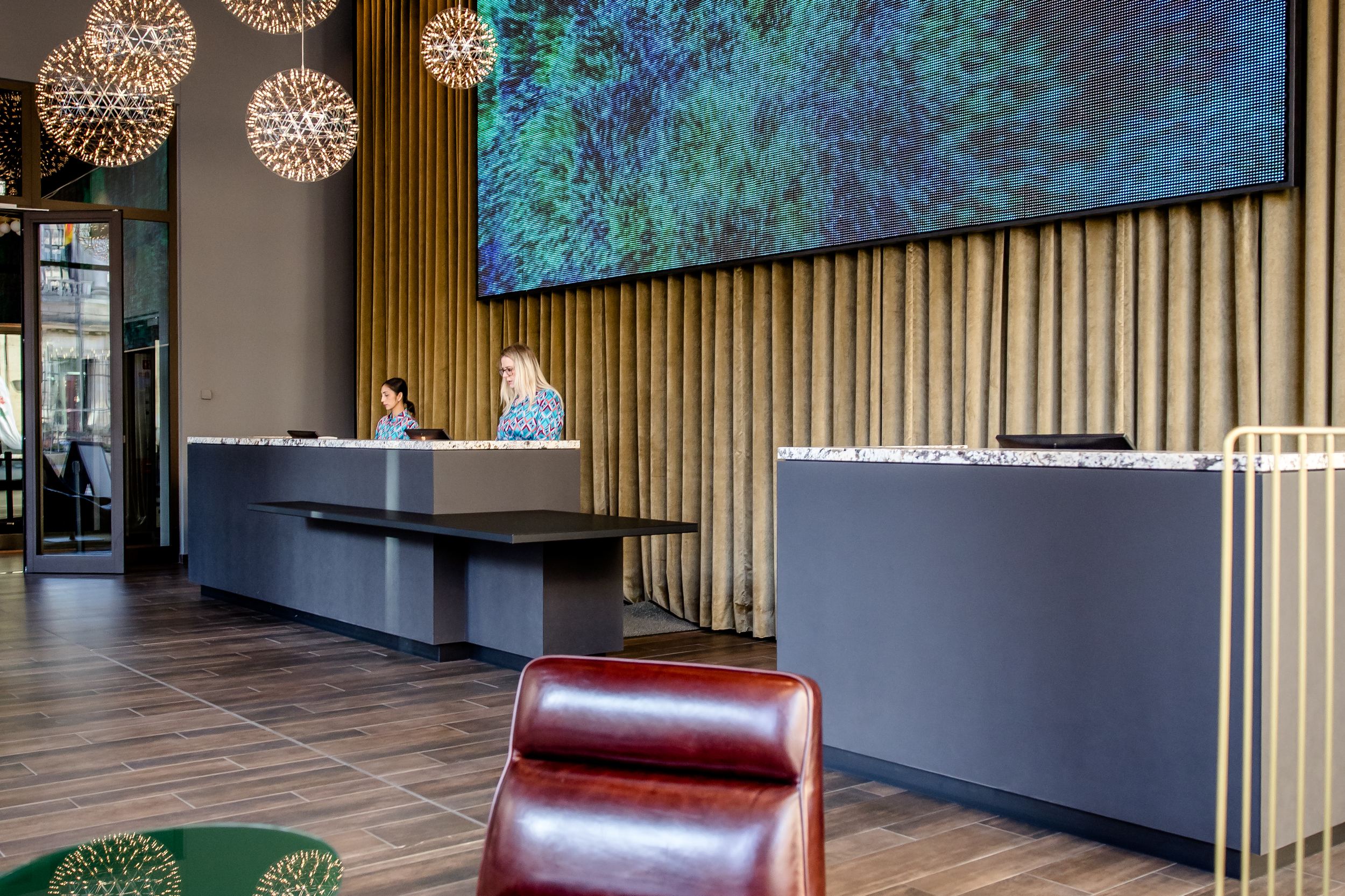 Hotel Berlin-Potsdamer Platz Motel One Rezeption mit eleganter Sitzecke, auffälliger Lichtinstallation aus kugelförmigen Leuchten und großem digitalem Kunstwerk an der Wand.