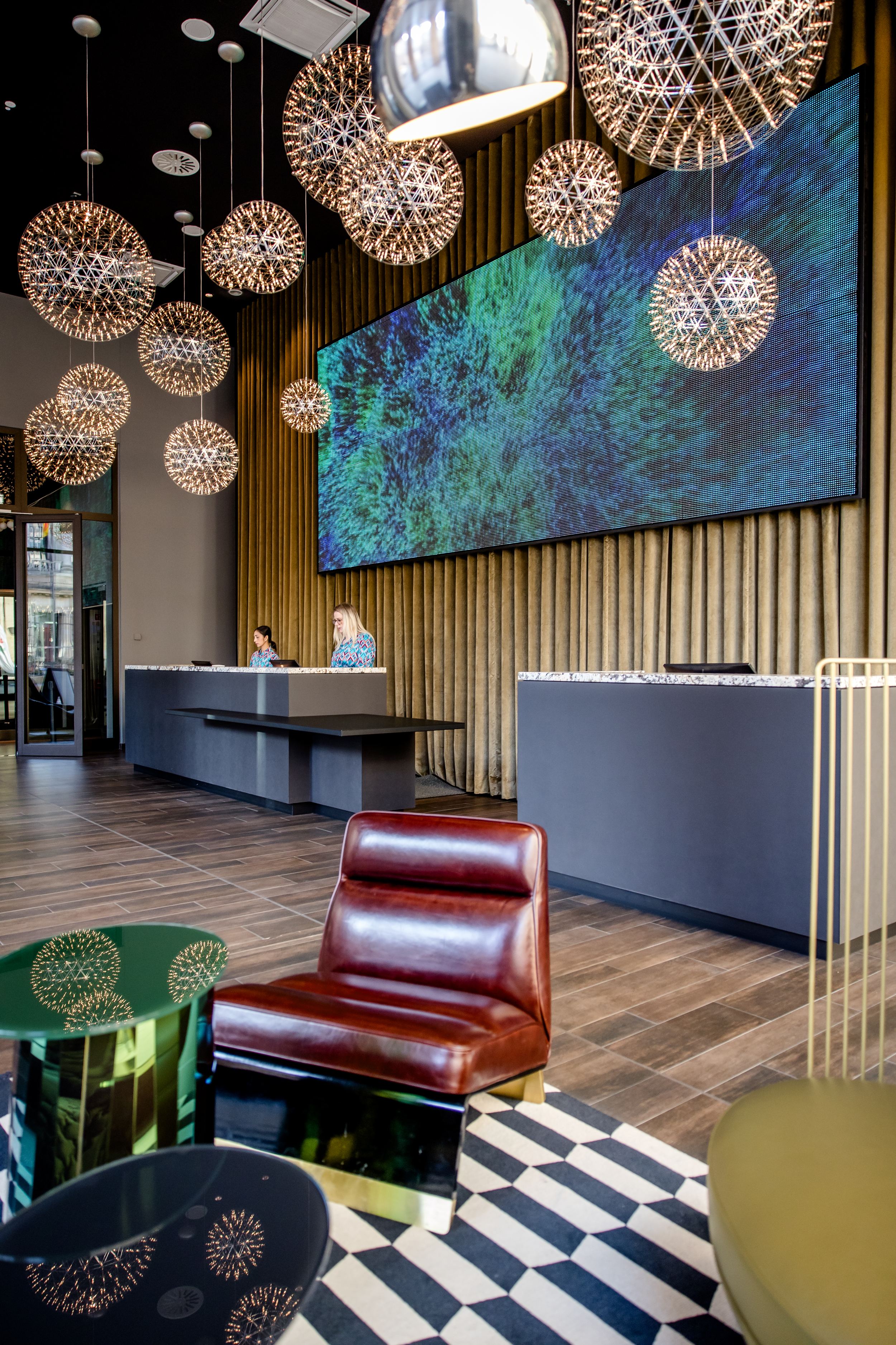 Hotel Berlin-Potsdamer Platz Motel One Rezeption mit eleganter Sitzecke, auffälliger Lichtinstallation aus kugelförmigen Leuchten und großem digitalem Kunstwerk an der Wand.