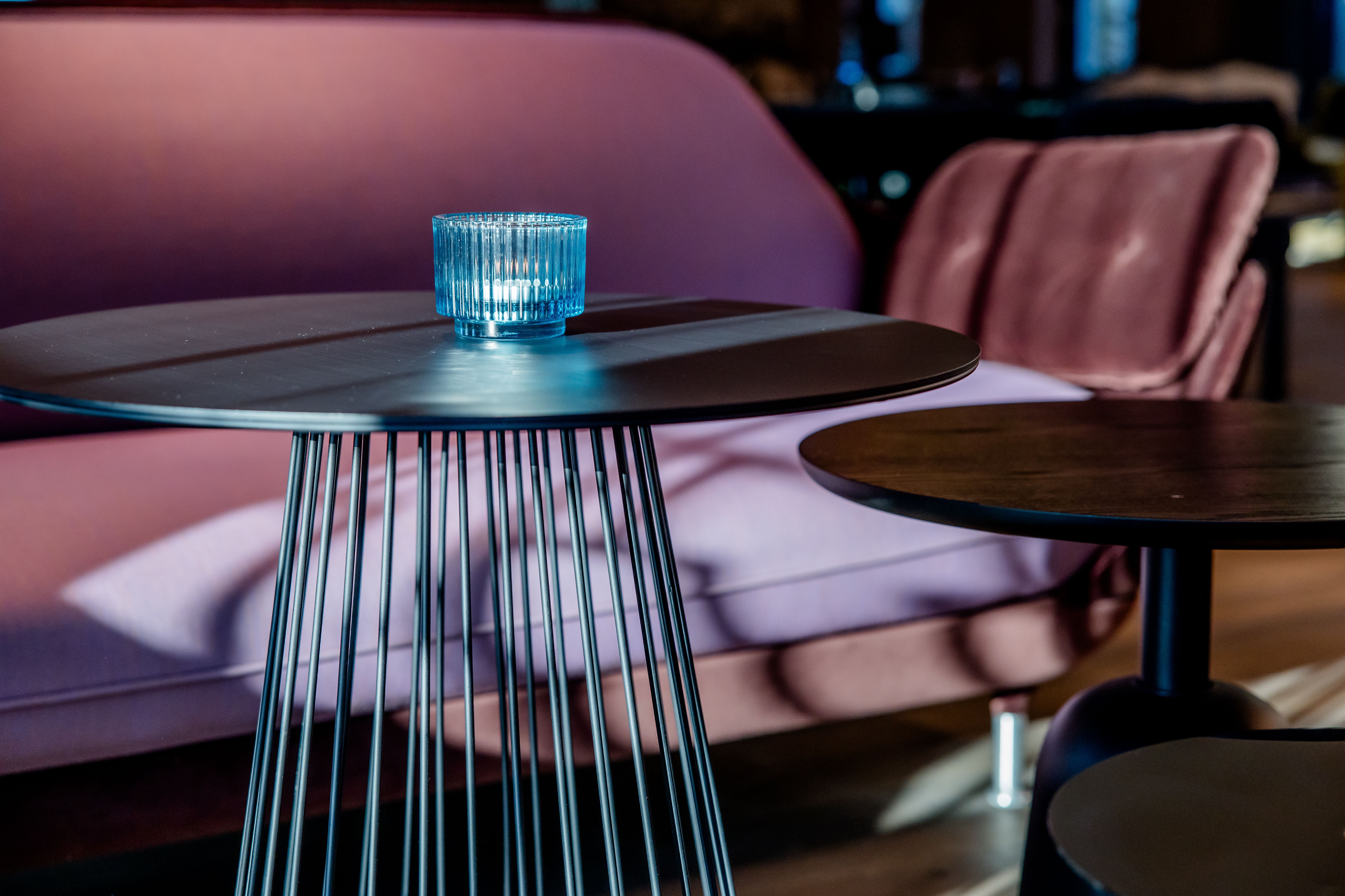 Designhotel Berlin-Potsdamer Platz Motel One Moderner Sitzbereich mit stilvollen, runden Metalltischen und einem rosa gepolsterten Sofa, auf einem Tisch steht ein blauer, dekorativer Kerzenhalter für eine gemütliche Atmosphäre.