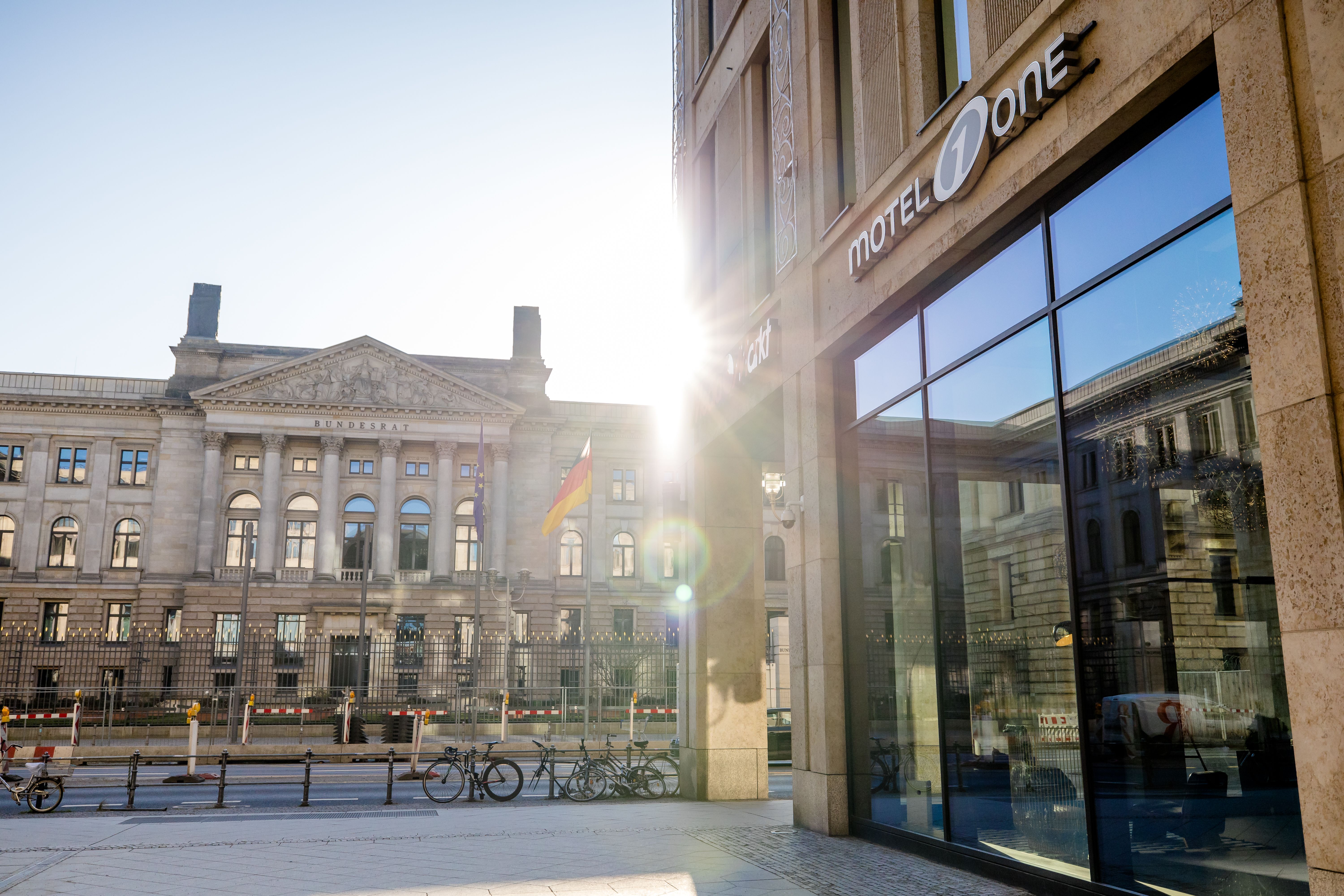 Hotel Berlin-Potsdamer Platz Motel One Fassade des modernen Motel One Hotels mit großer Glasfront, im Hintergrund ein historisches Regierungsgebäude mit deutscher Flagge bei Sonnenlicht. 