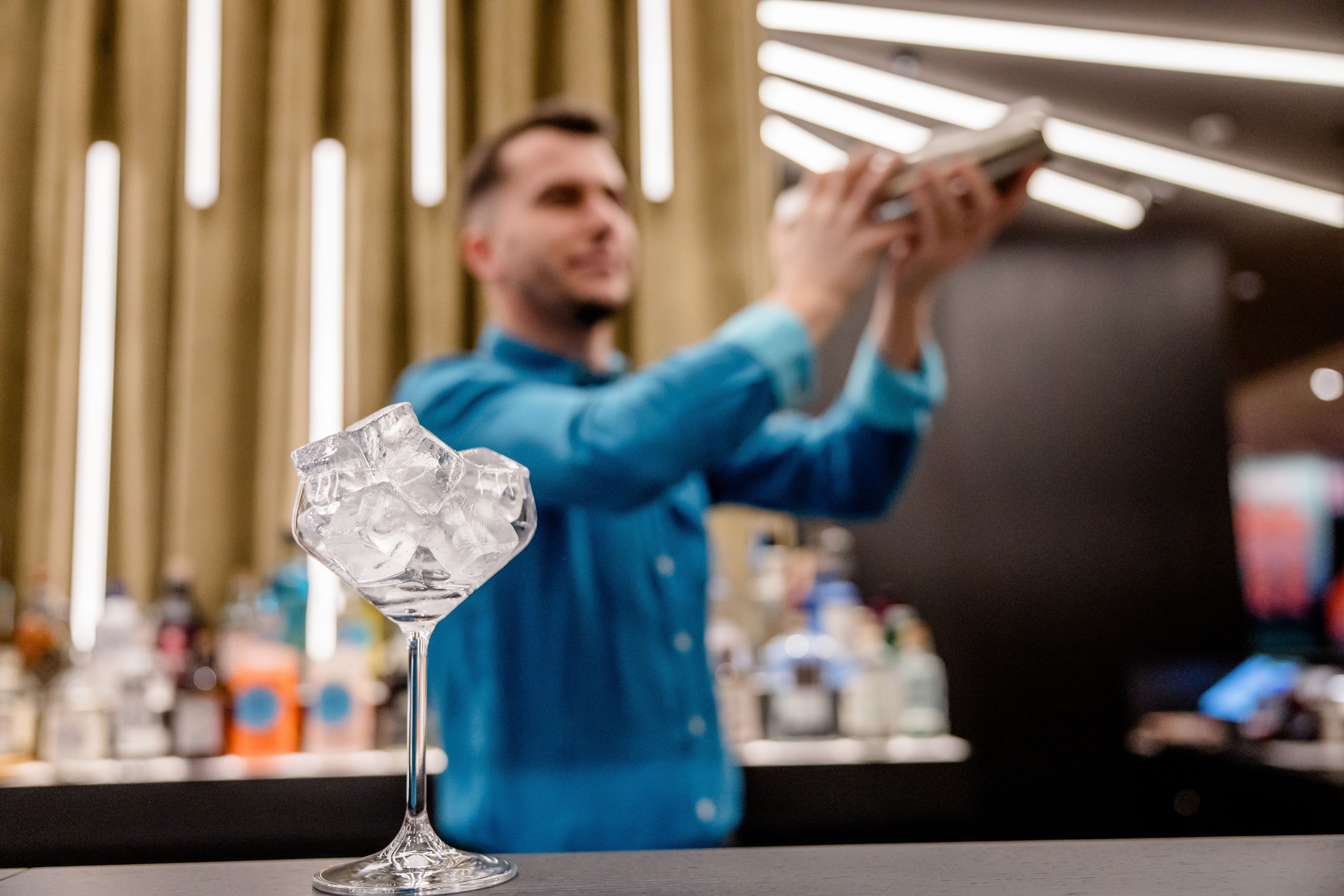 Hotel Berlin-Potsdamer Platz Motel One Barkeeper schüttelt Cocktailshaker hinter Bar, im Vordergrund ist ein mit Eis gefülltes Cocktailglas auf der Theke zu sehen.