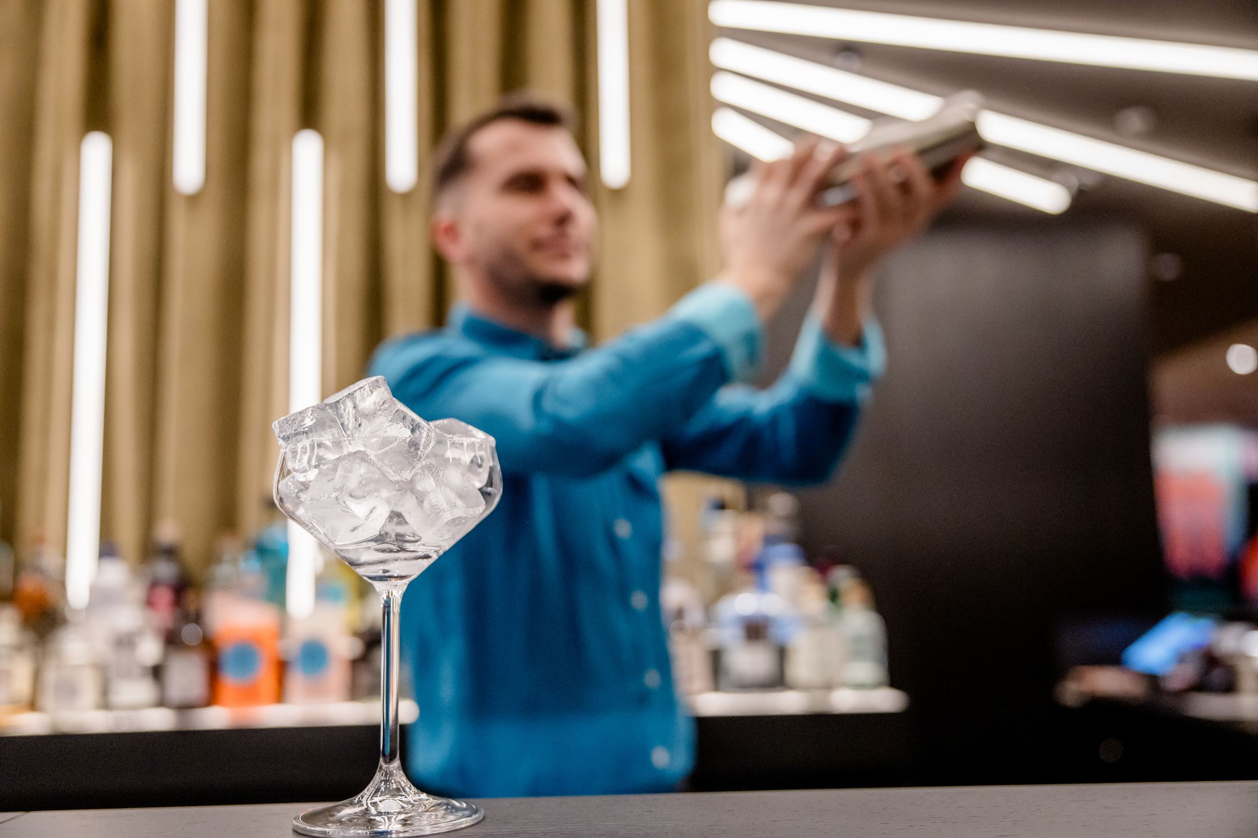 Hotel Berlin-Potsdamer Platz Motel One Bartender shakes cocktail shaker behind bar; cocktail glass filled with ice can be seen on counter in foreground.