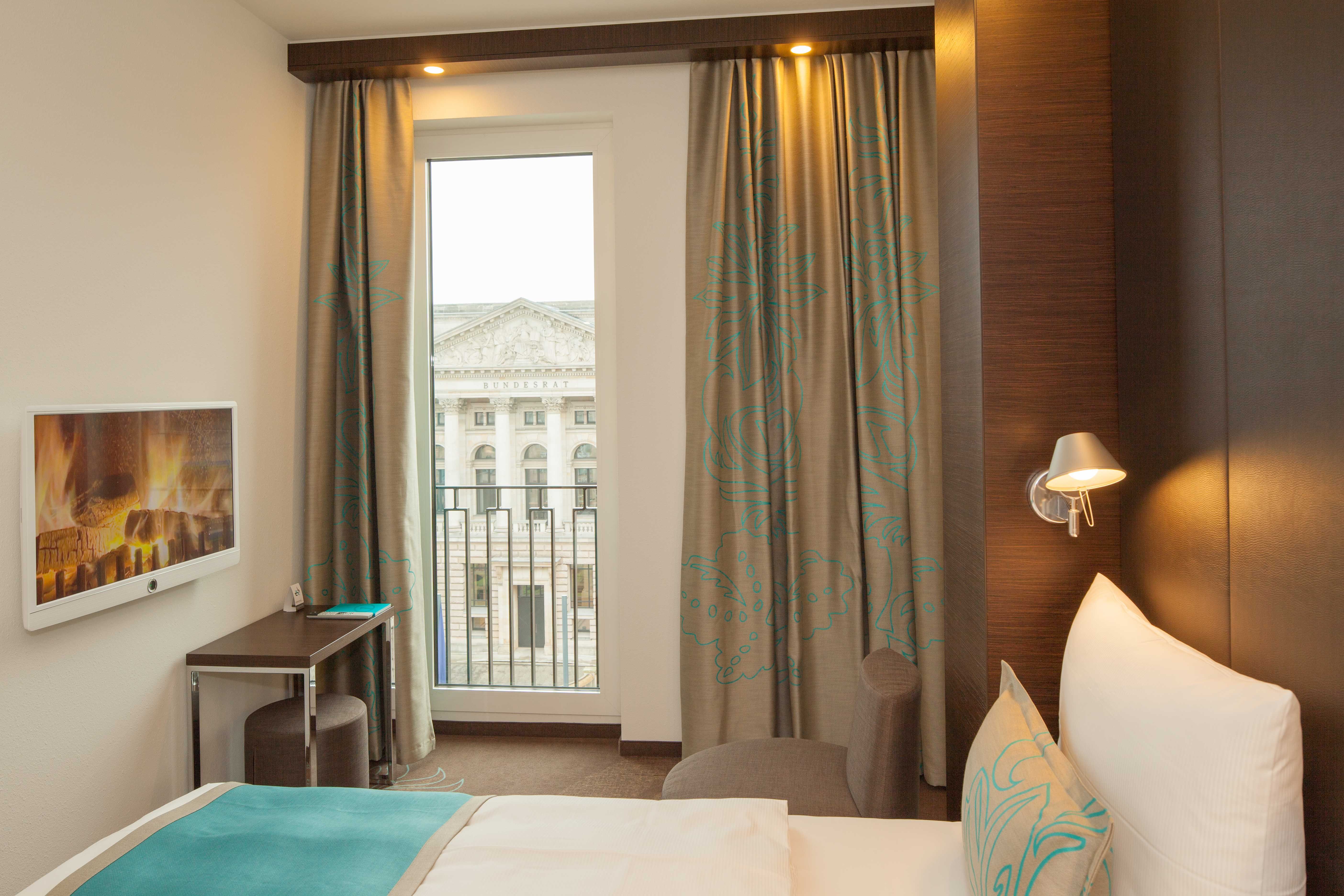 Hotel Berlin Potsdamer Platz Motel One helles Zimmer mit großem Bett, Fernseher an der Wand sowie Hocker und Tisch vor bodentiefem Fenster