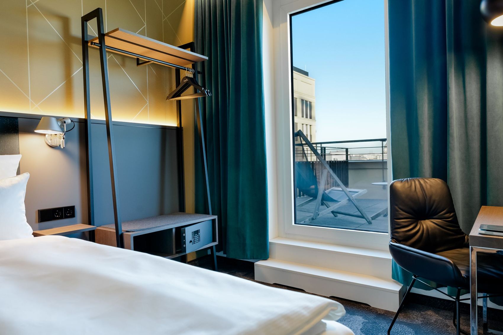 Hotel Berlin-Potsdamer Platz Motel One - Modern eingerichtetes Hotelzimmer mit großem Fenster und Zugang zu einer Terrasse, komfortables Bett, Schreibtisch mit Stuhl, offene Garderobe und beruhigende Farbgestaltung in Blau- und Grautönen.
