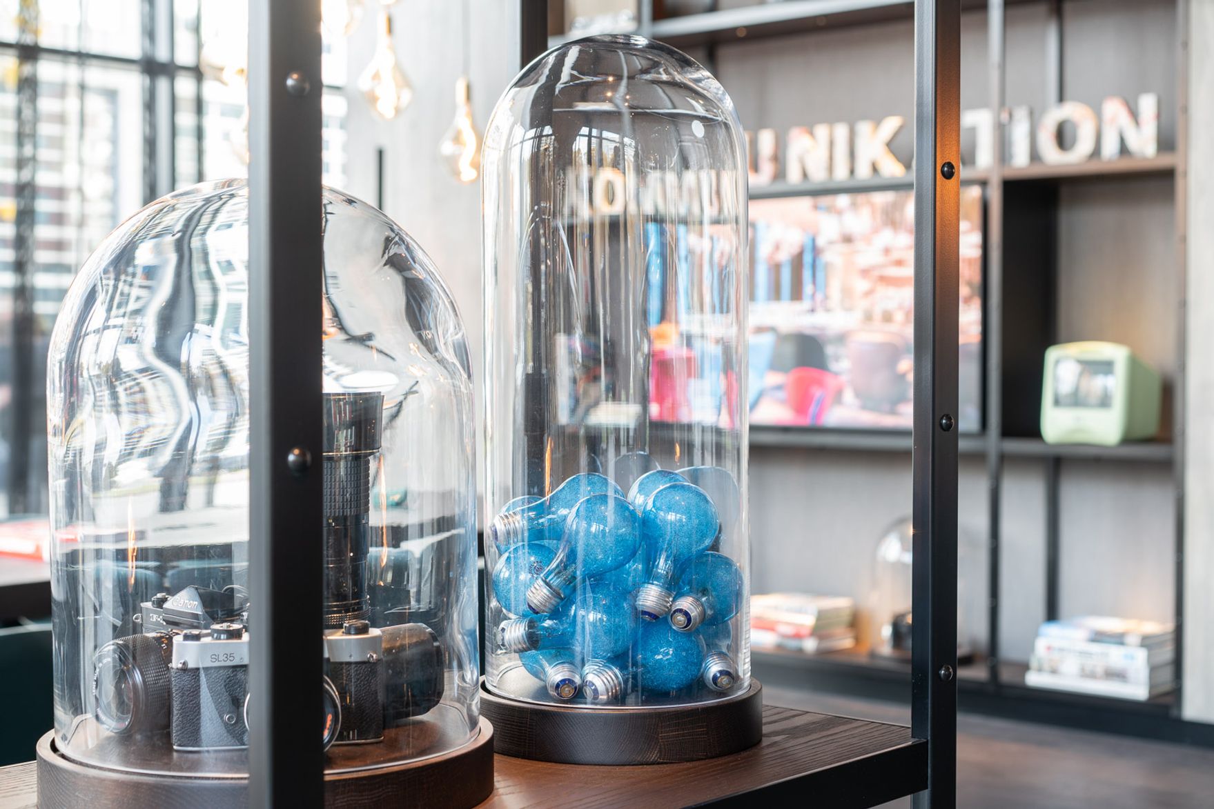 Designhotel Berlin Spittelmarkt Motel One Nahaufnahme auf ein offenes Regal mit blauen Glühbirnen unter einer Glasglocke sowie Kameras unter einer weiteren Glasglocke