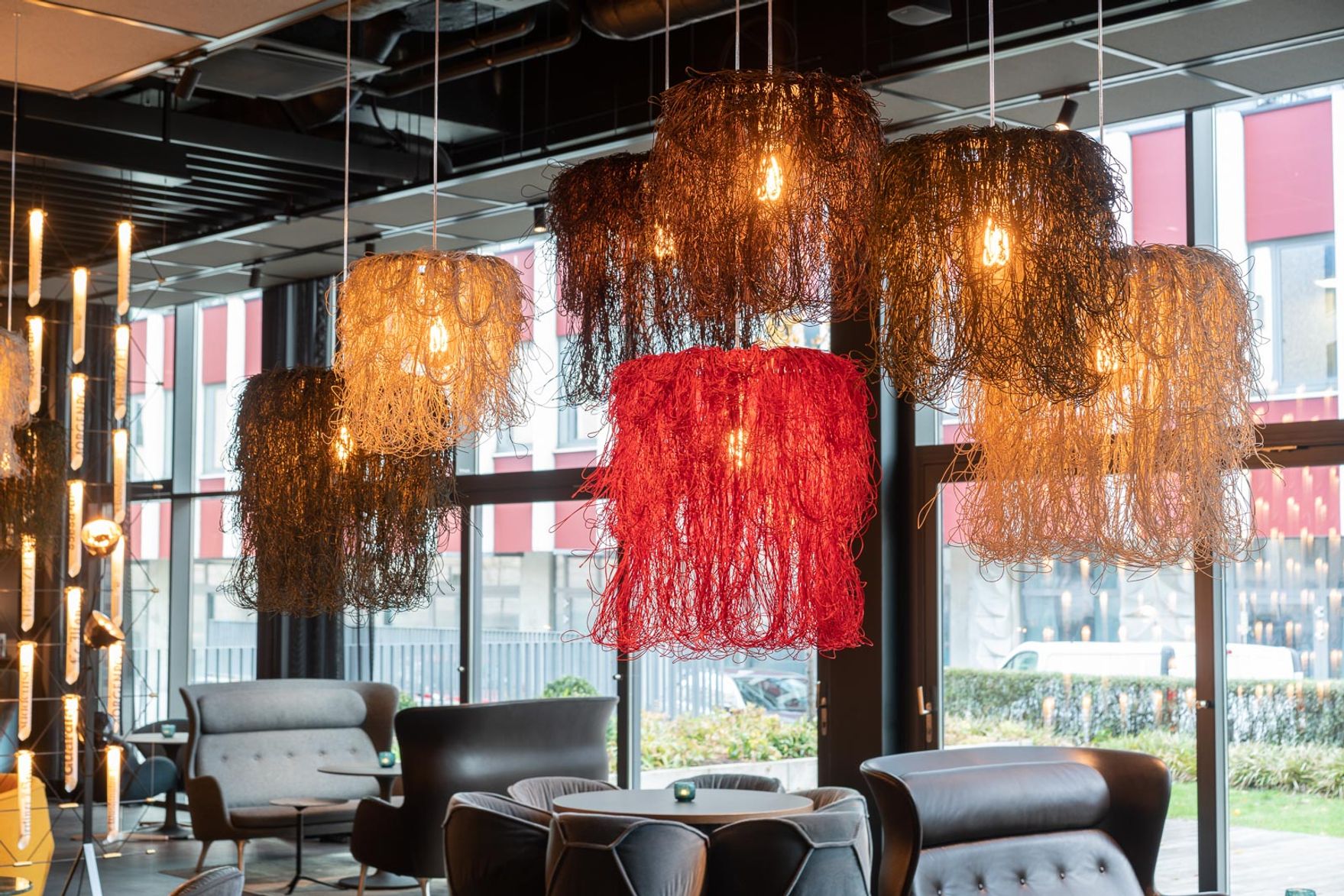 Designhotel Berlin Spittelmarkt Motel One Lounge mit bodentiefer Fensterfront und mehreren Sesseln sowie Blick auf rote, gelbe und braune Lampenschirme, die von der Decke hängen