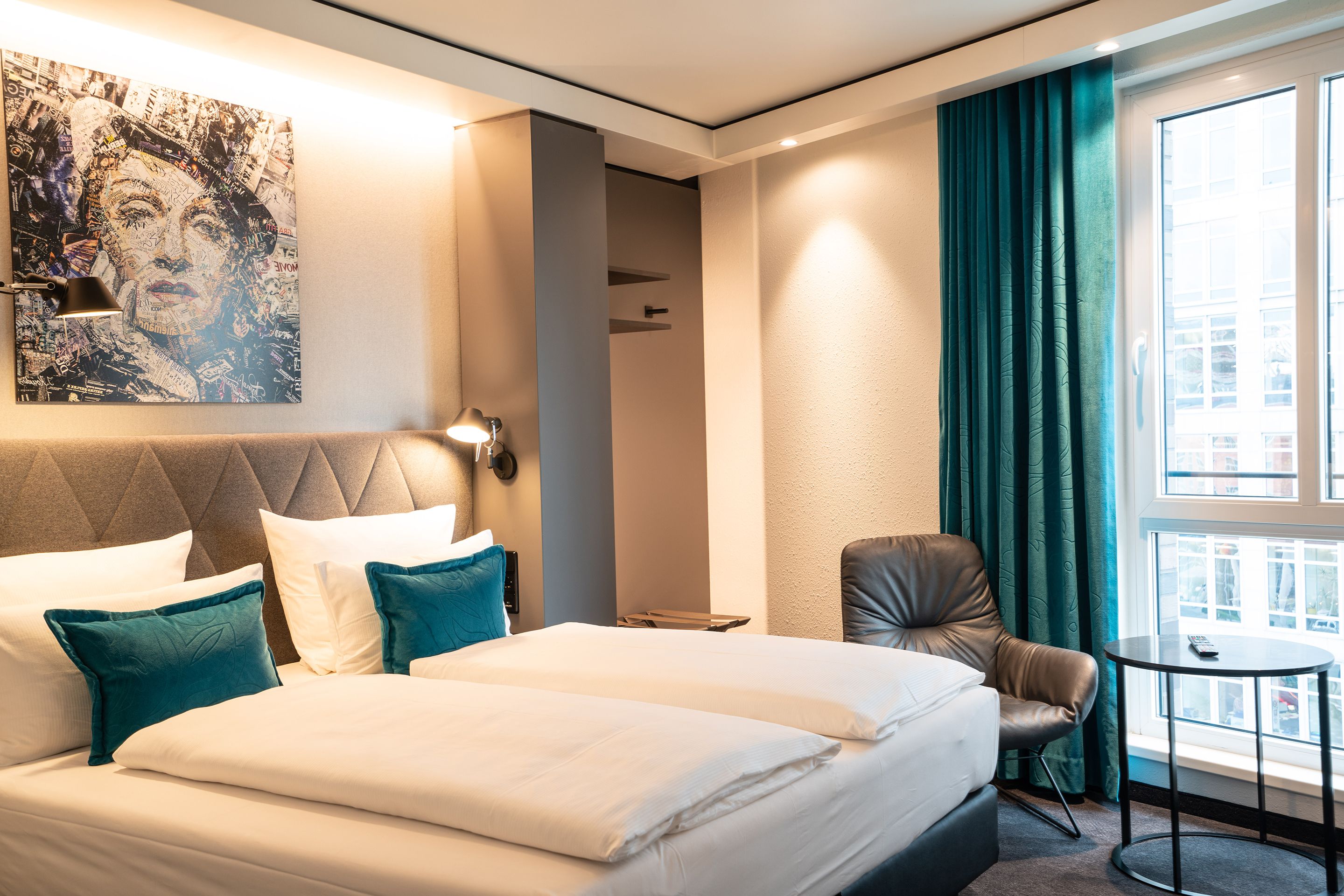 Hotel Motel One Berlin-Spittelmarkt Modernes Hotelzimmer mit großem Doppelbett, türkisfarbenen Kissen, gemütlichem Sessel, bodentiefen Fenstern und dunklen Vorhängen, wärmer Beleuchtung und dekorativem Kunstbild über dem Bett.