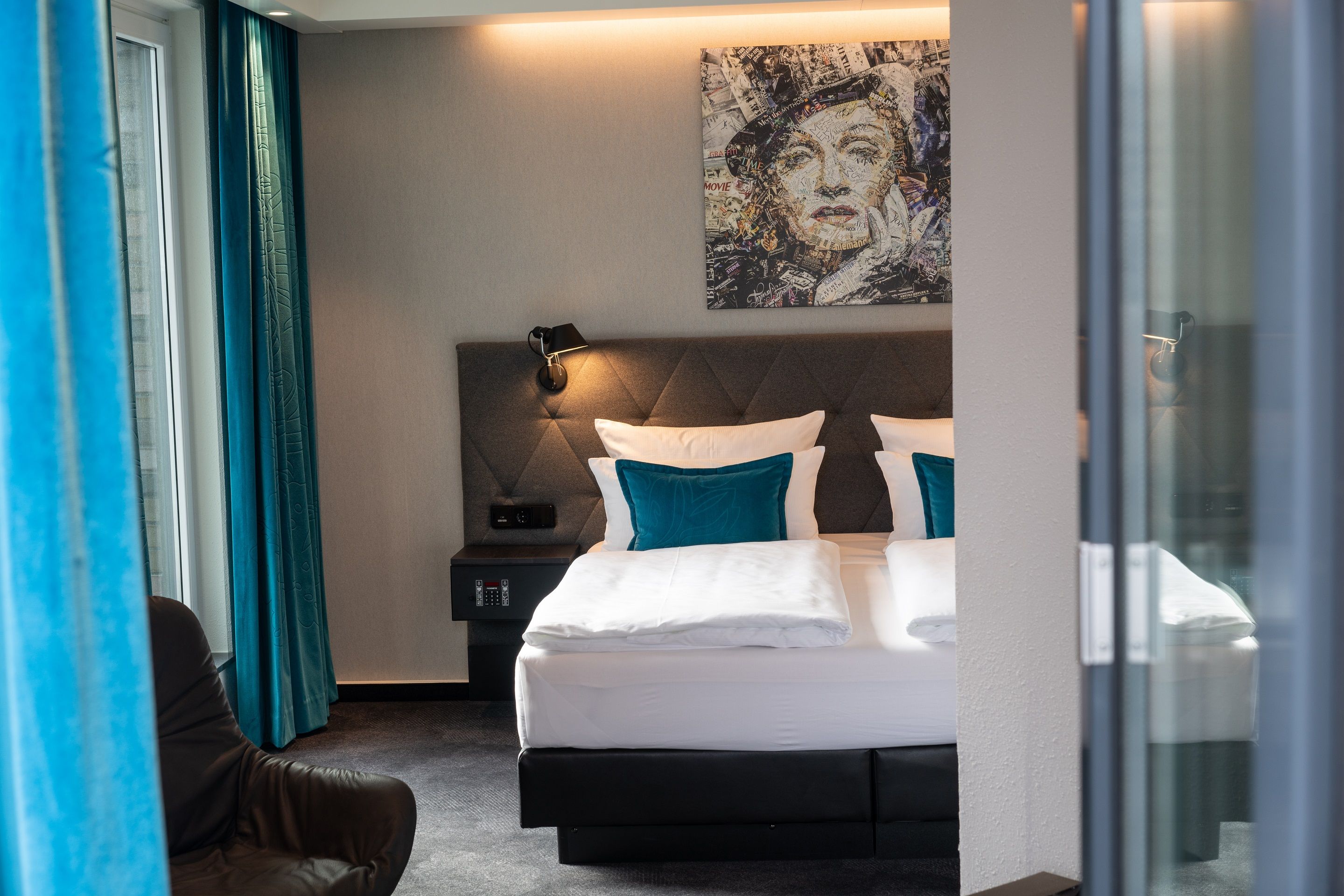 Hotel Motel One Berlin-Spittelmarkt Modernes Hotelzimmer mit komfortablem Doppelbett, blauen Akzentkissen, eleganten Vorhängen und stimmungsvoller Beleuchtung. An der Wand hängt ein künstlerisches Porträt.