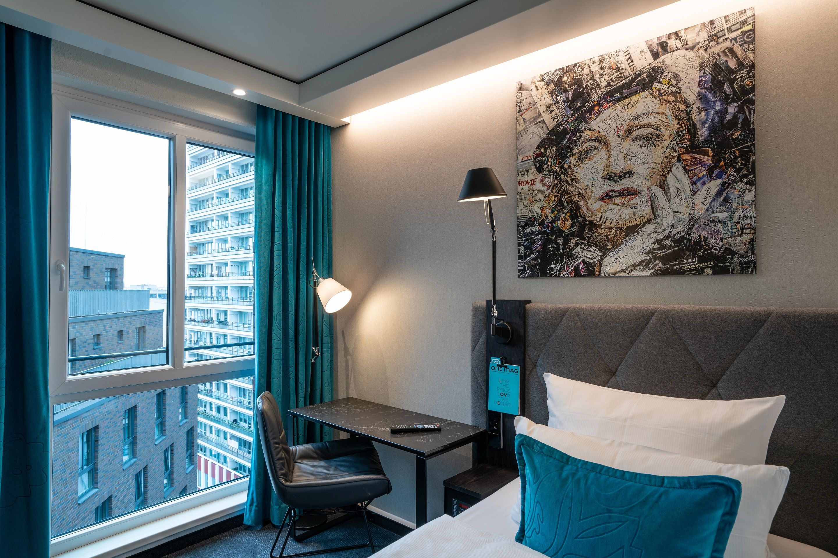 Hotel Motel One Berlin-Spittelmarkt Gemütliches Hotelzimmer mit großem Fensterausblick auf Hochhäuser, moderner Schreibtischecke, stilvoller Beleuchtung und einem Bett mit weißen Kissen und blauem Dekokissen. An der Wand hängt ein künstlerisches Porträt.