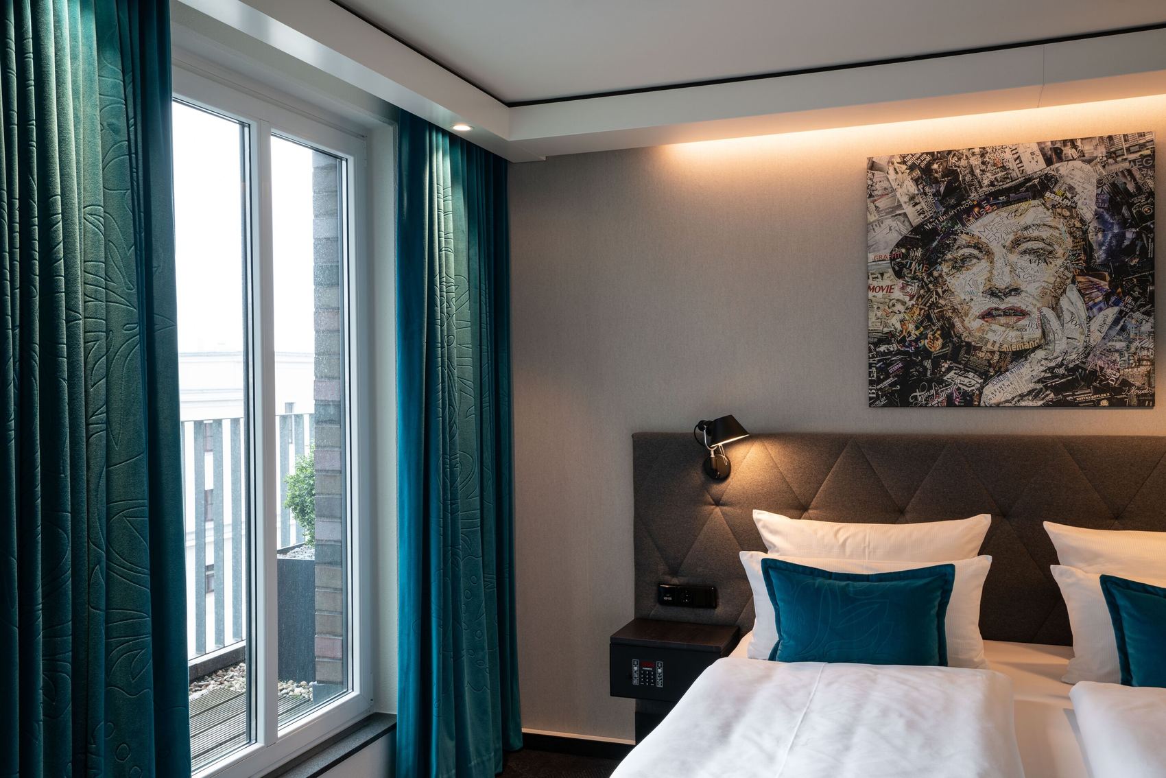 Hotel Motel One Berlin-Spittelmarkt Modernes Hotelzimmer mit großem Fenster und Balkon, gemütlichem Doppelbett mit blauen Akzentkissen, stimmungsvoller Beleuchtung und stilvollem Kunstwerk über dem Kopfteil.