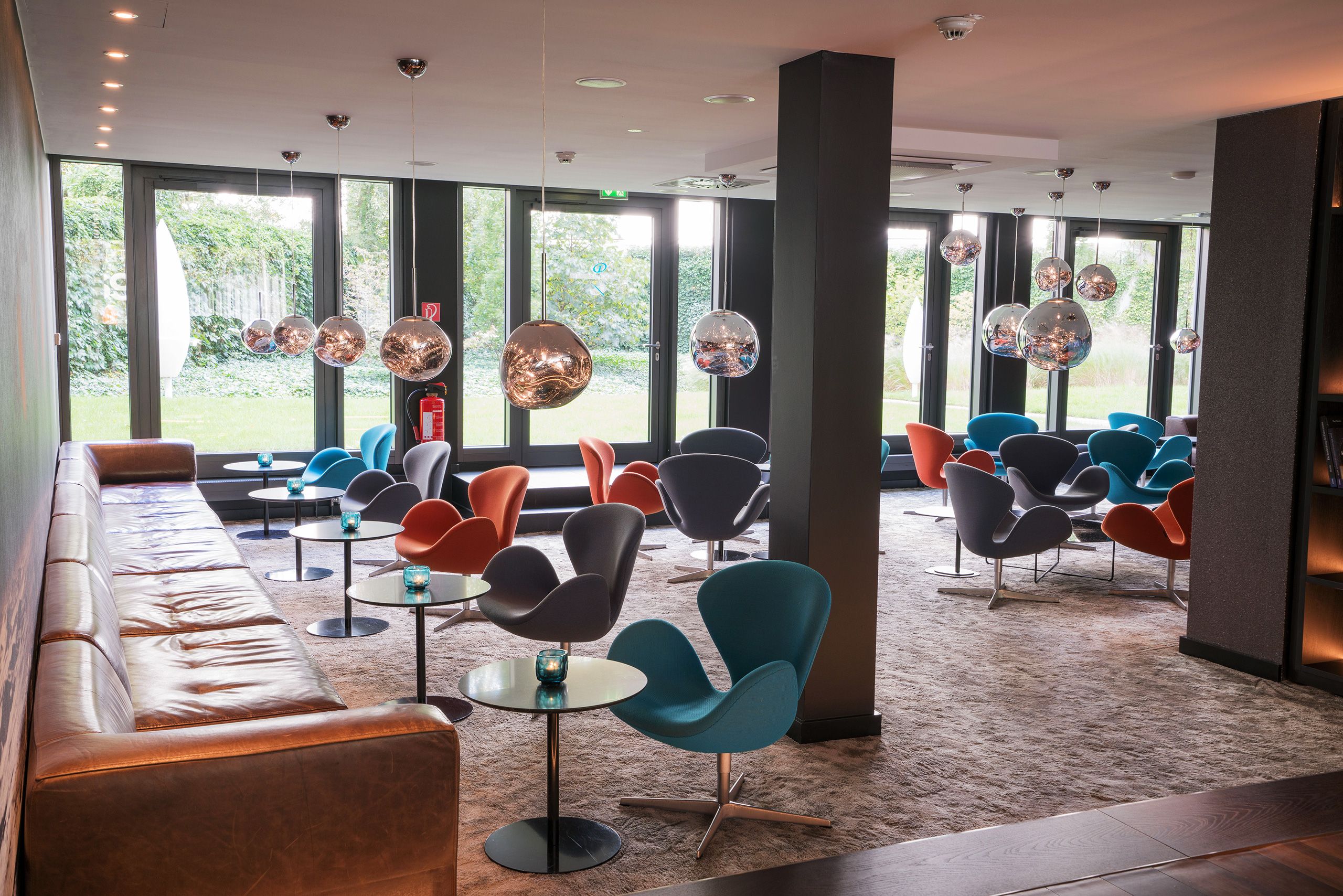 Hotel Berlin Tiergarten Motel One helle Lounge mit Sofas, Sesseln und Tischen sowie mehrere runde Lampen in silber und bodentiefe Fenster mit Blick ins Grüne