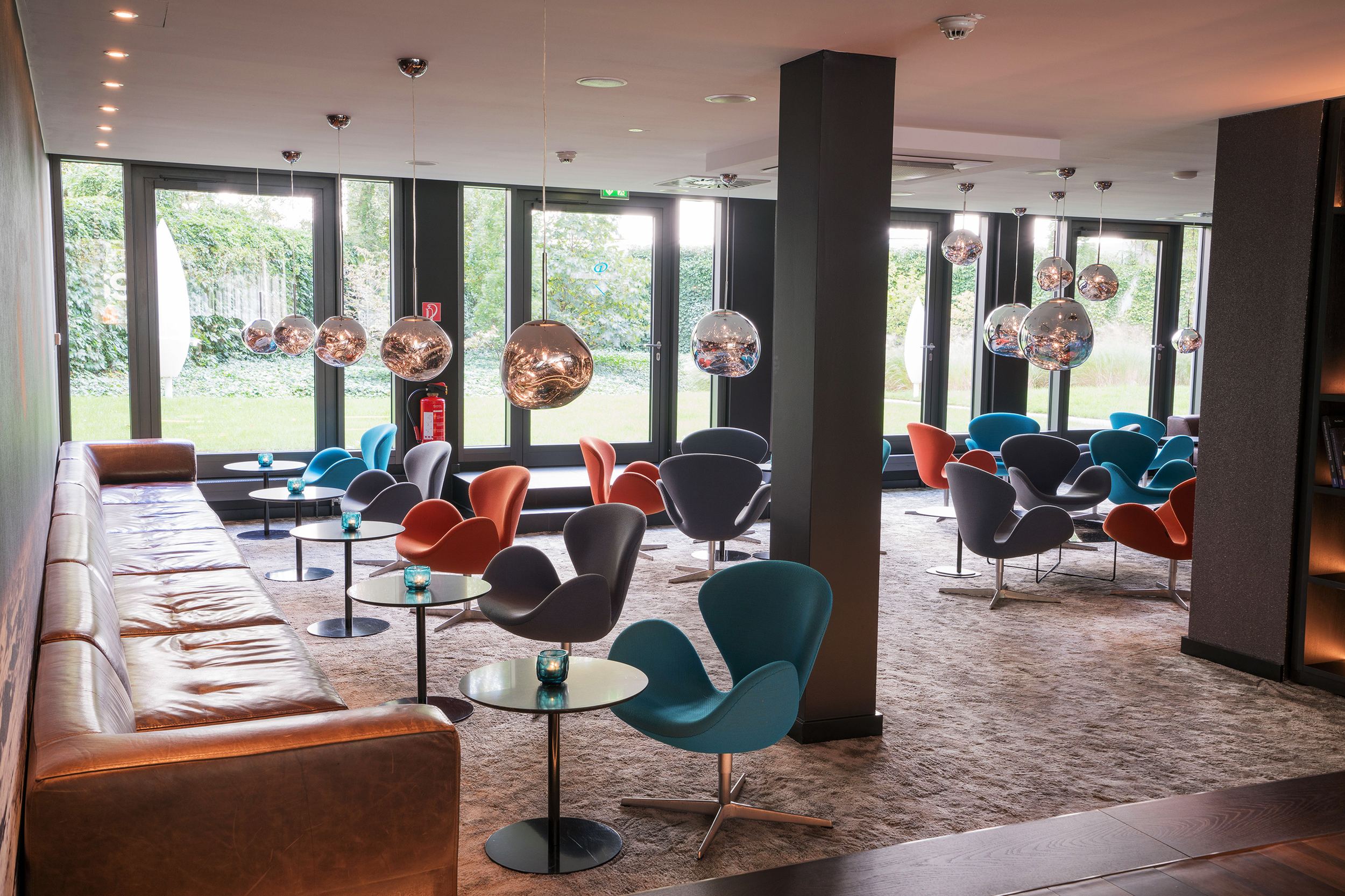 hotel Berlin Tiergarten Motel One bright lounge with sofas, armchairs, and tables, as well as several round silver lamps and floor-to-ceiling windows with a view of the greenery