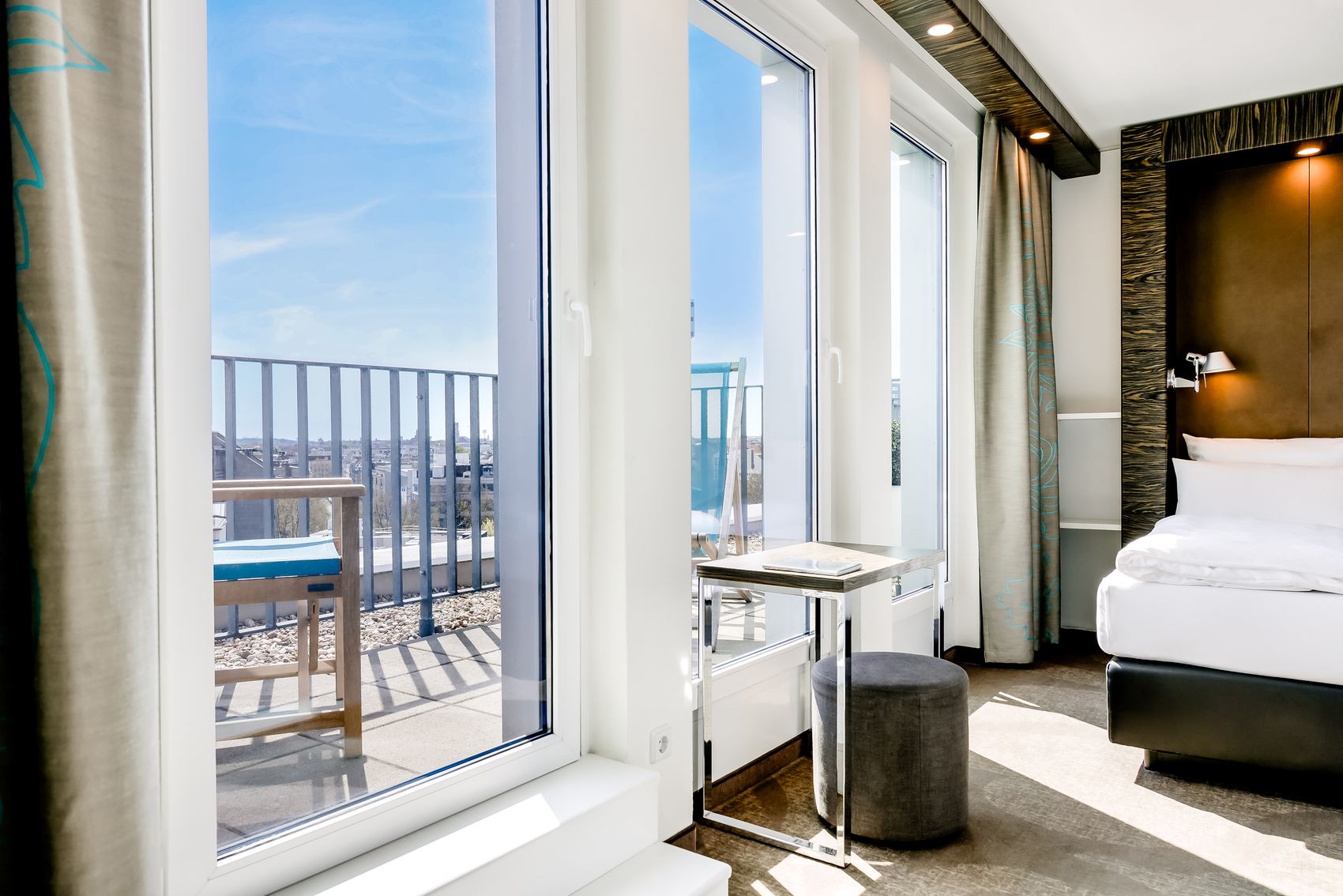 Hell eingerichtetes Hotelzimmer mit großem Fenster und Zugang zu einem Balkon. Blick auf die Stadt und blauen Himmel. Komfortables Bett und Sitzgelegenheit am Fenster schaffen eine einladende, entspannte Atmosphäre für Gäste.