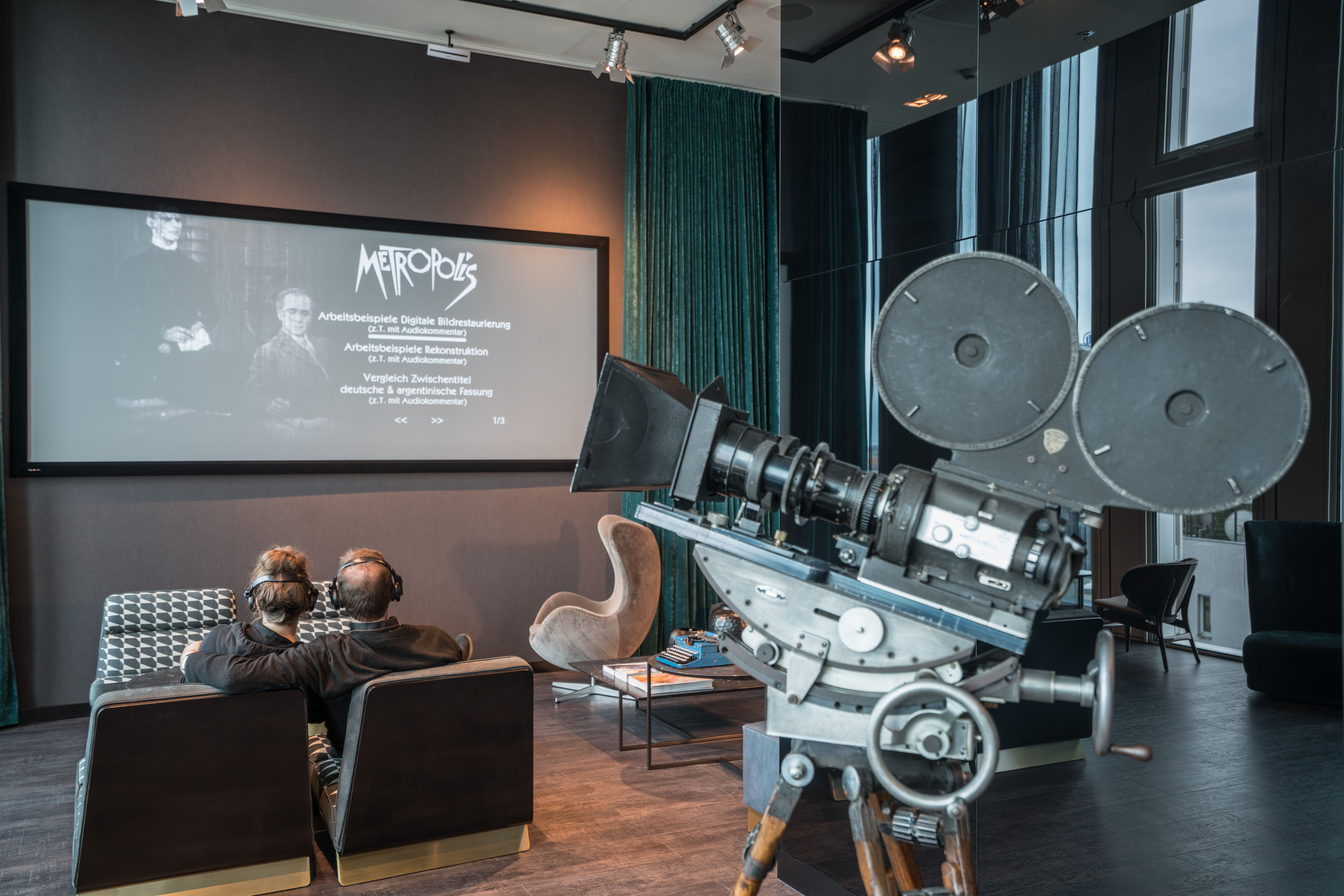 Designhotel Berlin Upper West Motel One Lounge mit zwei Personen, die mit großen Kopfhörern auf einem Sofa sitzen und auf eine Leinwand mit Film schauen sowie eine Filmkamera im Vordergrund