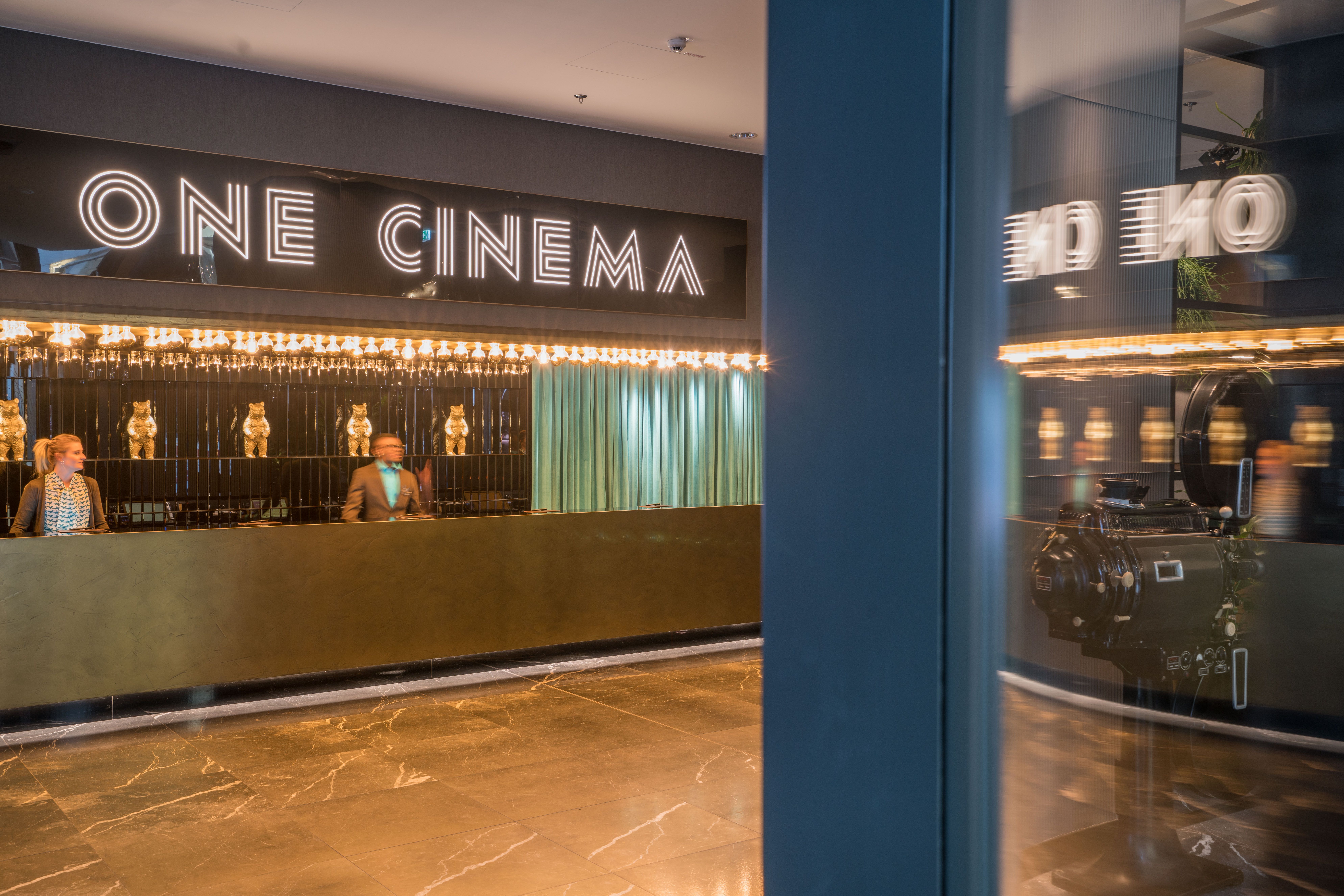 Designhotel Berlin Upper West Motel One Rezeption mit Mitarbeitenden bei der Arbeit sowie einem großen Schriftzug "One Cinema" über der Rezeption
