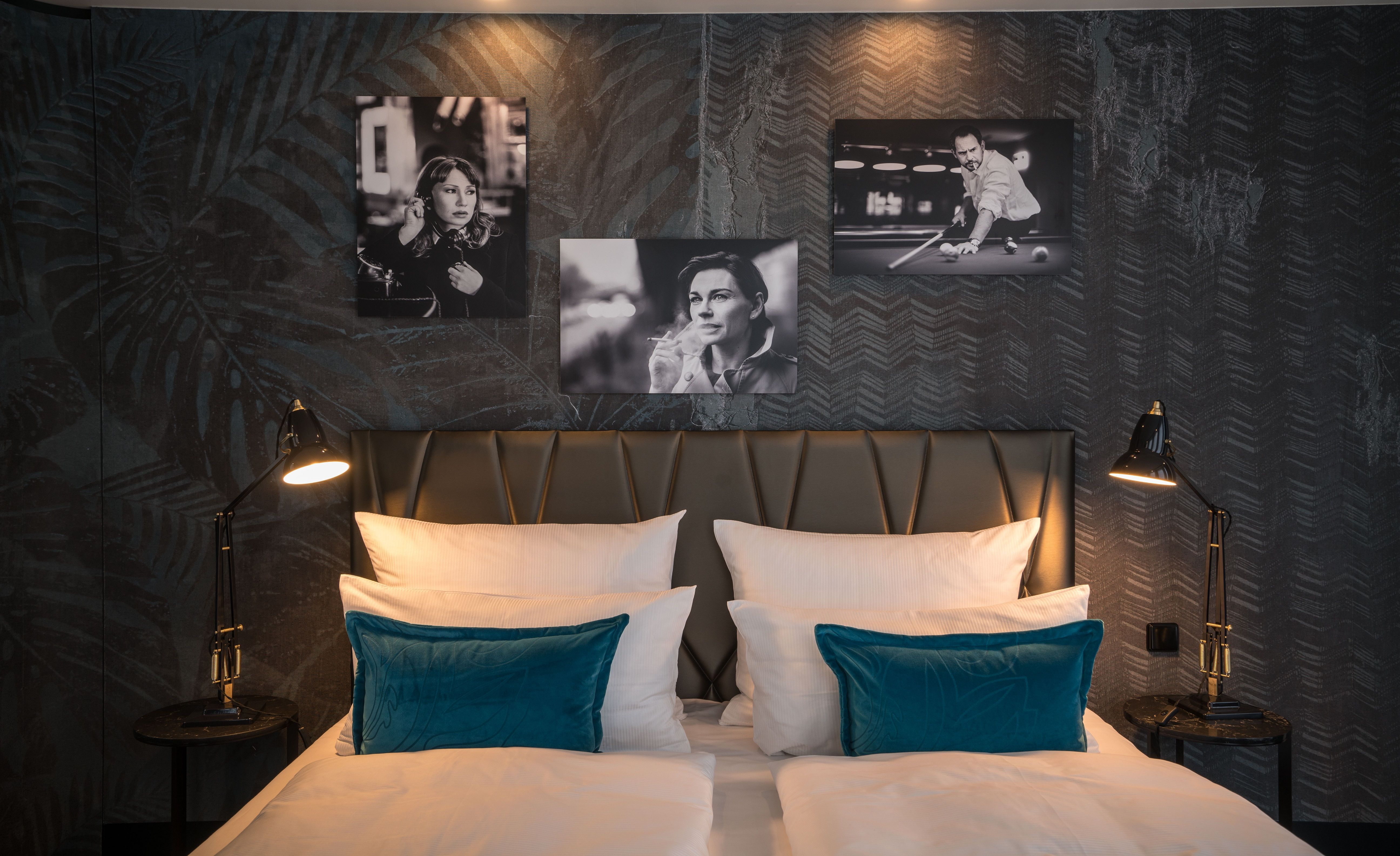 Designhotel Berlin Upper West Motel One Zimmer mit Blick auf ein Doppelbett mit Zierkissen in türkis sowie drei Bilder in schwarz-weiß von Schauspielern an der Wand