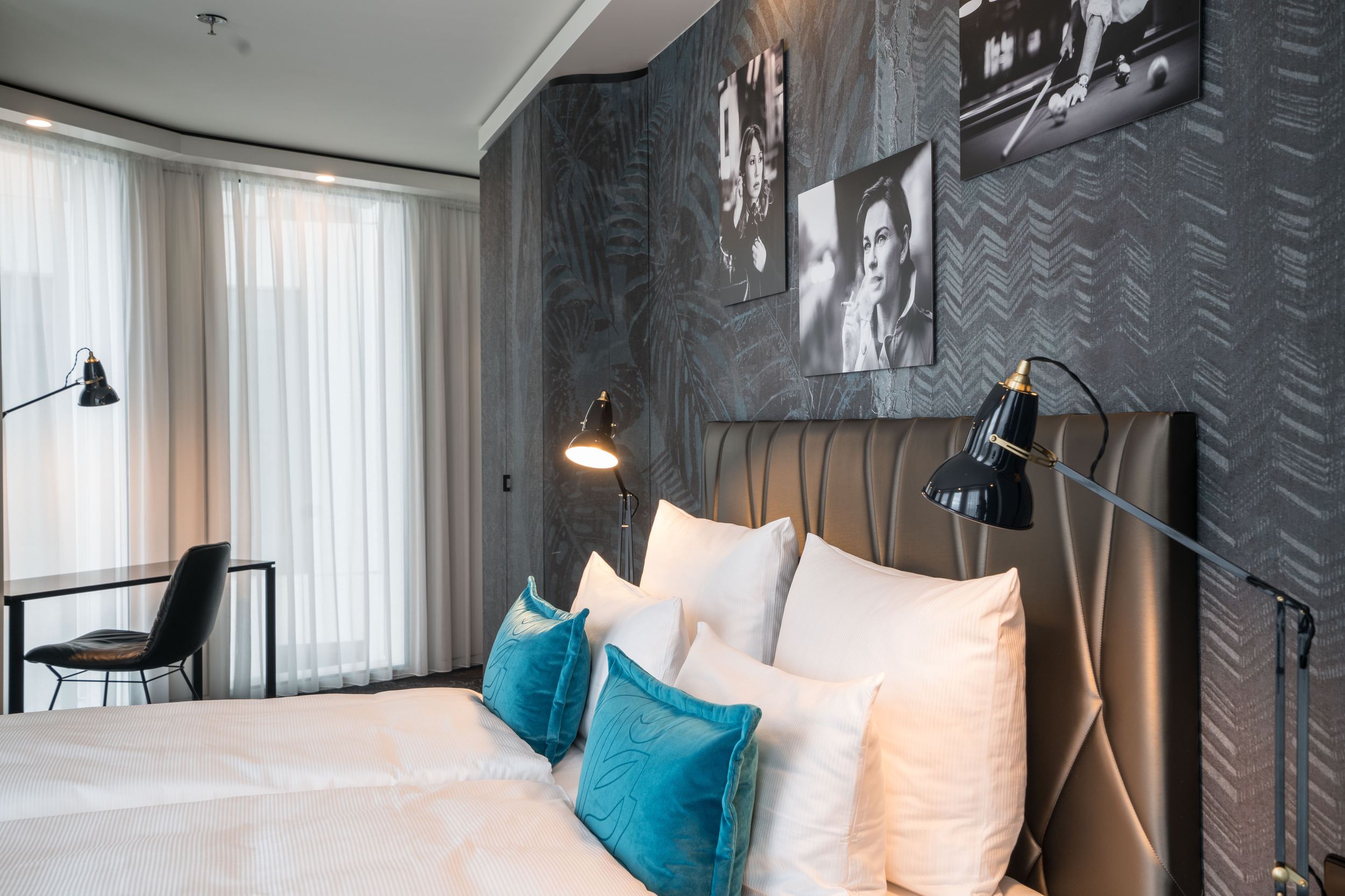 Designhotel Berlin Upper West Motel One seitlicher Blick auf ein Doppelbett mit Zierkissen in türkis sowie Bilder in schwarz-weiß von Schauspielern an der Wand und ein Schreibtisch mit Stuhl vor bodentiefem Fenster