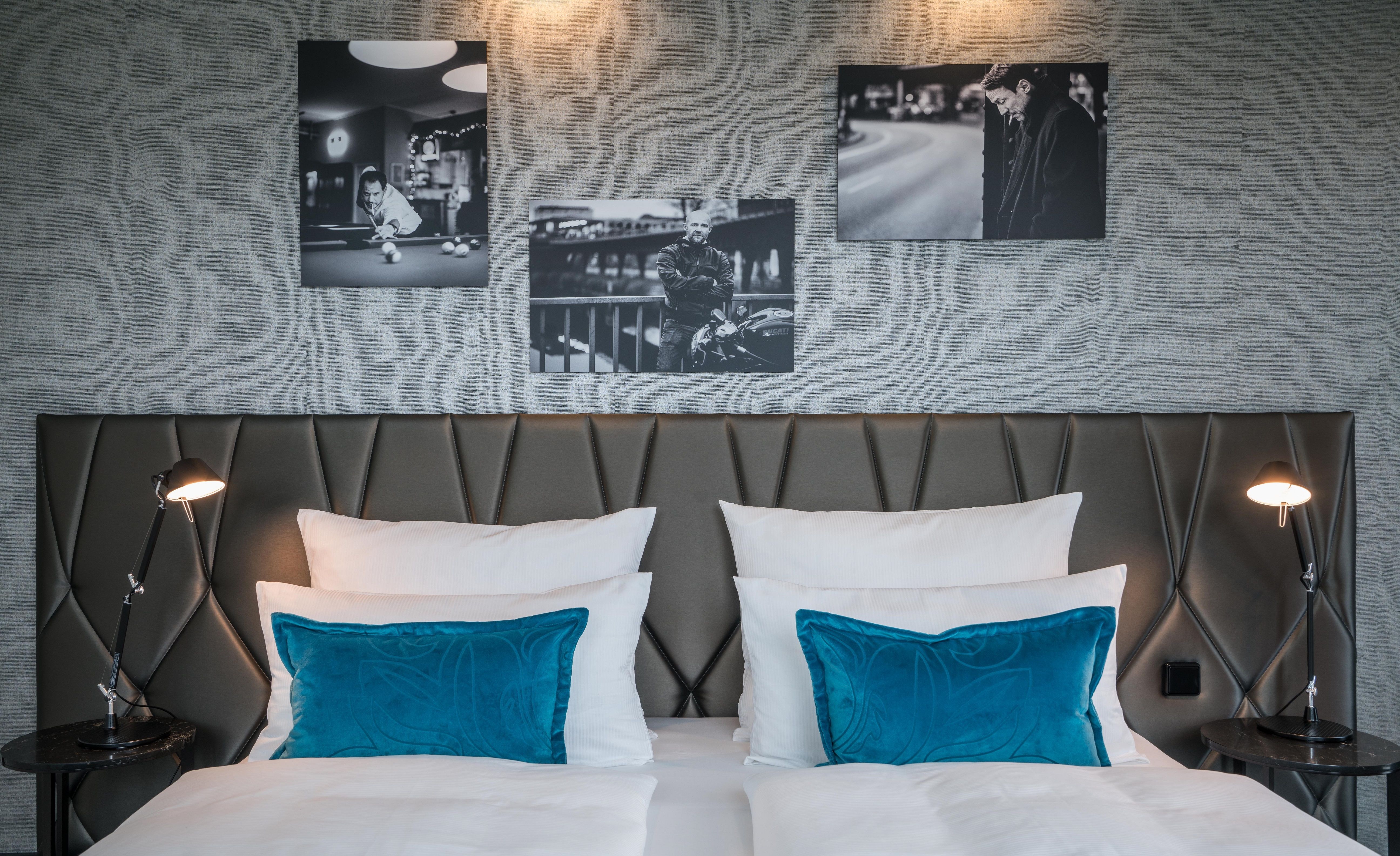 Designhotel Berlin Upper West Motel One Blick auf ein großes Doppelbett mit weißen Bezügen und Zierkissen in türkis sowie drei Gemälde an der Wand