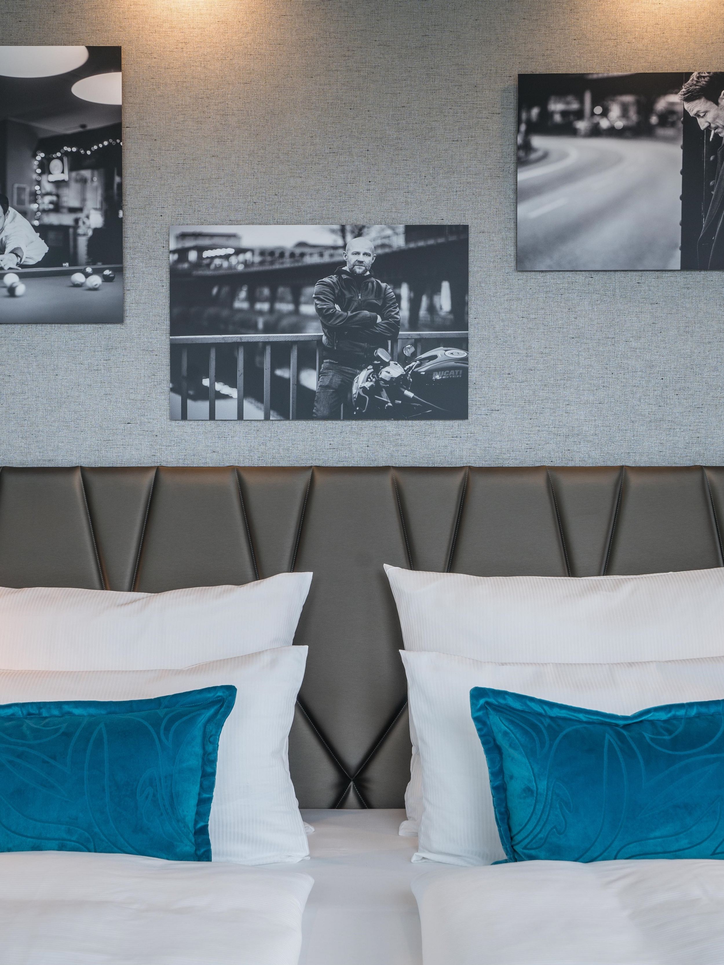 Designhotel Berlin Upper West Motel One Blick auf ein großes Doppelbett mit weißen Bezügen und Zierkissen in türkis sowie drei Gemälde an der Wand