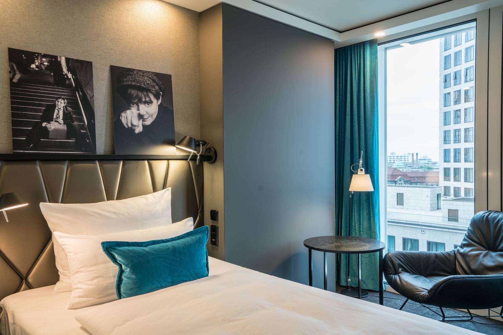 Designhotel Berlin Upper West Motel One helles Zimmer mit Einzelbett, Sessel mit Beistelltisch vor großem Fenster sowie zwei Bilder von Schauspielern in schwarz-weiß an der Wand über dem Bett