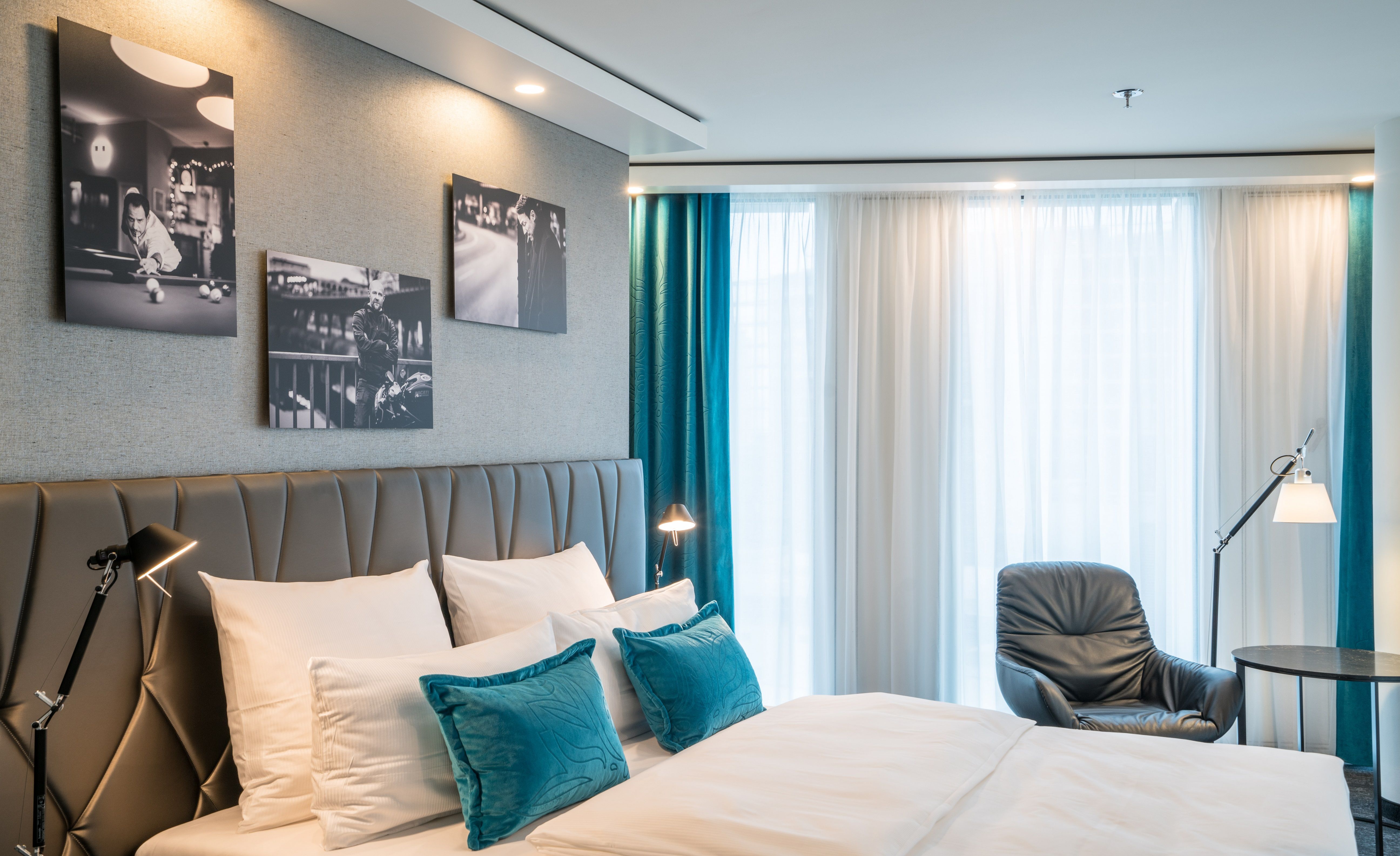 Designhotel Berlin Upper West Motel One seitlicher Blick in ein helles Zimmer mit komfortablem Doppelbett, einem Stuhl mit Tisch und Stehlampe vor großer Fensterfront mit Vorhängen sowie drei Bilder an der Wand