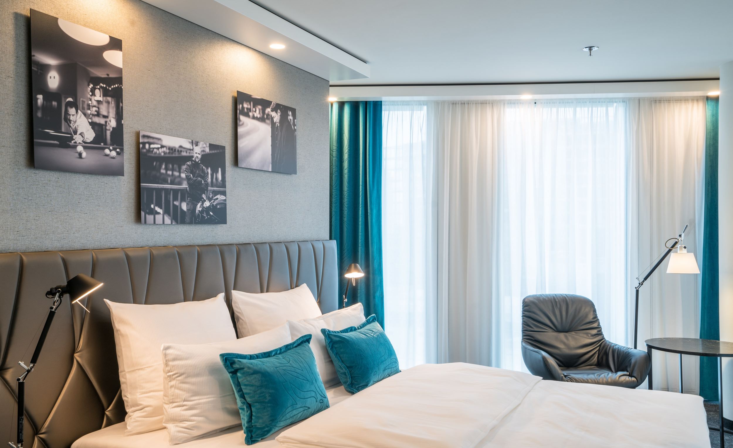 Designhotel Berlin Upper West Motel One seitlicher Blick in ein helles Zimmer mit komfortablem Doppelbett, einem Stuhl mit Tisch und Stehlampe vor großer Fensterfront mit Vorhängen sowie drei Bilder an der Wand